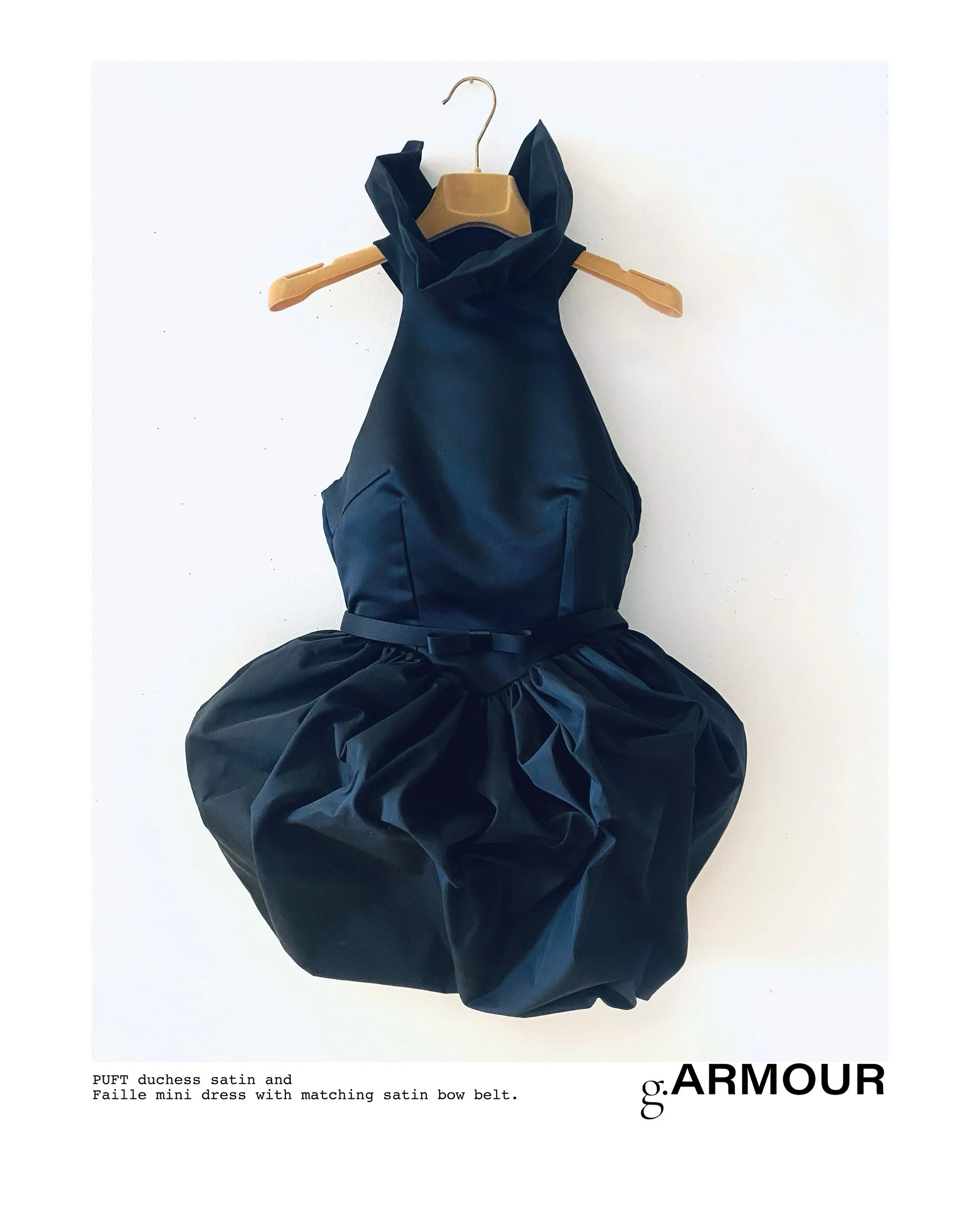 A black mini dress with a satin finish and matching satin belt on a wooden hanger against a white background.