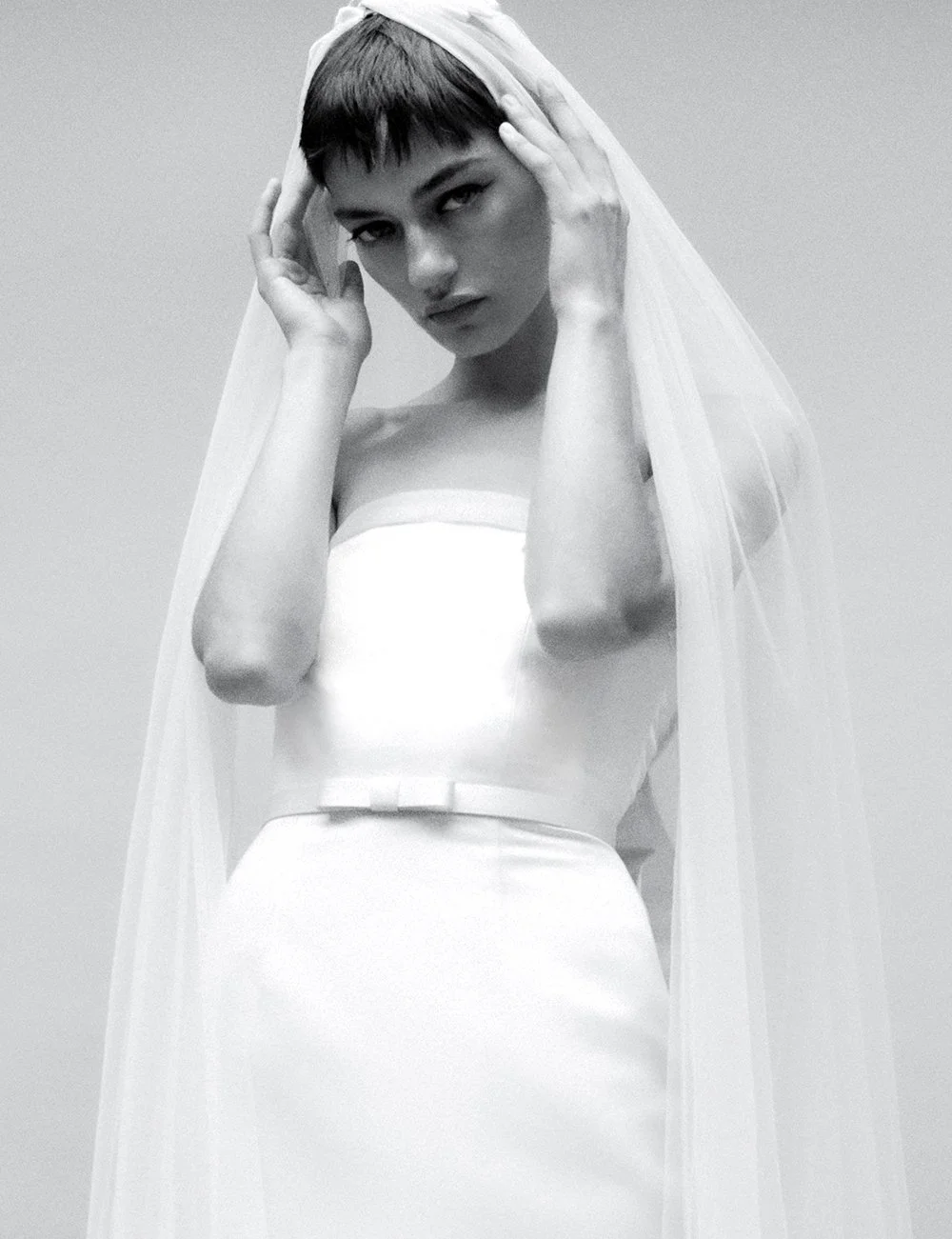 A black-and-white portrait of a woman with short hair, wearing a strapless dress and a sheer veil, holding the veil with both hands, looking directly at the camera with a serious expression.