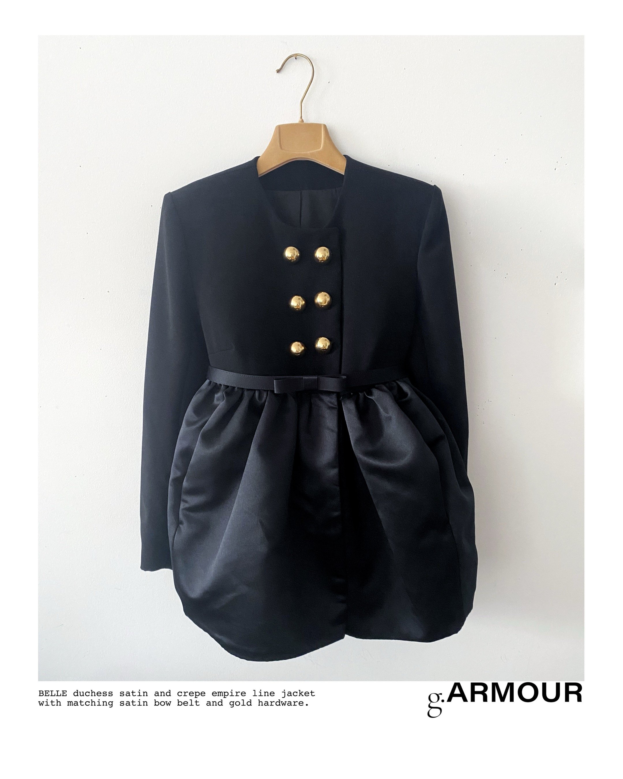 Black satin and crepe empire line jacket with gold hardware and matching satin bow belt, hanging on a hanger against a white wall.