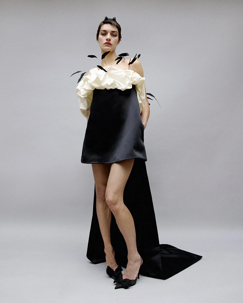 A woman in a black dress with a high slit, high heels, and black and white accessories, posing against a plain grey background.