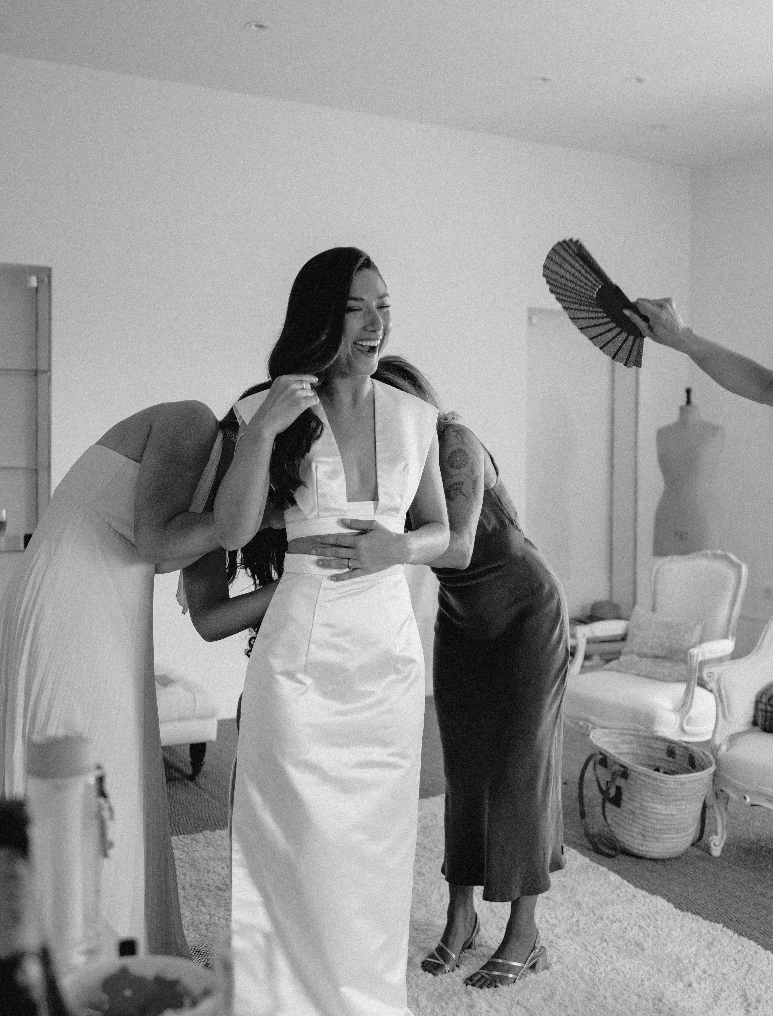 A woman smiling and laughing, wearing a white dress, surrounded by friends helping her get ready in a room with vintage furniture and a dress form in the background.