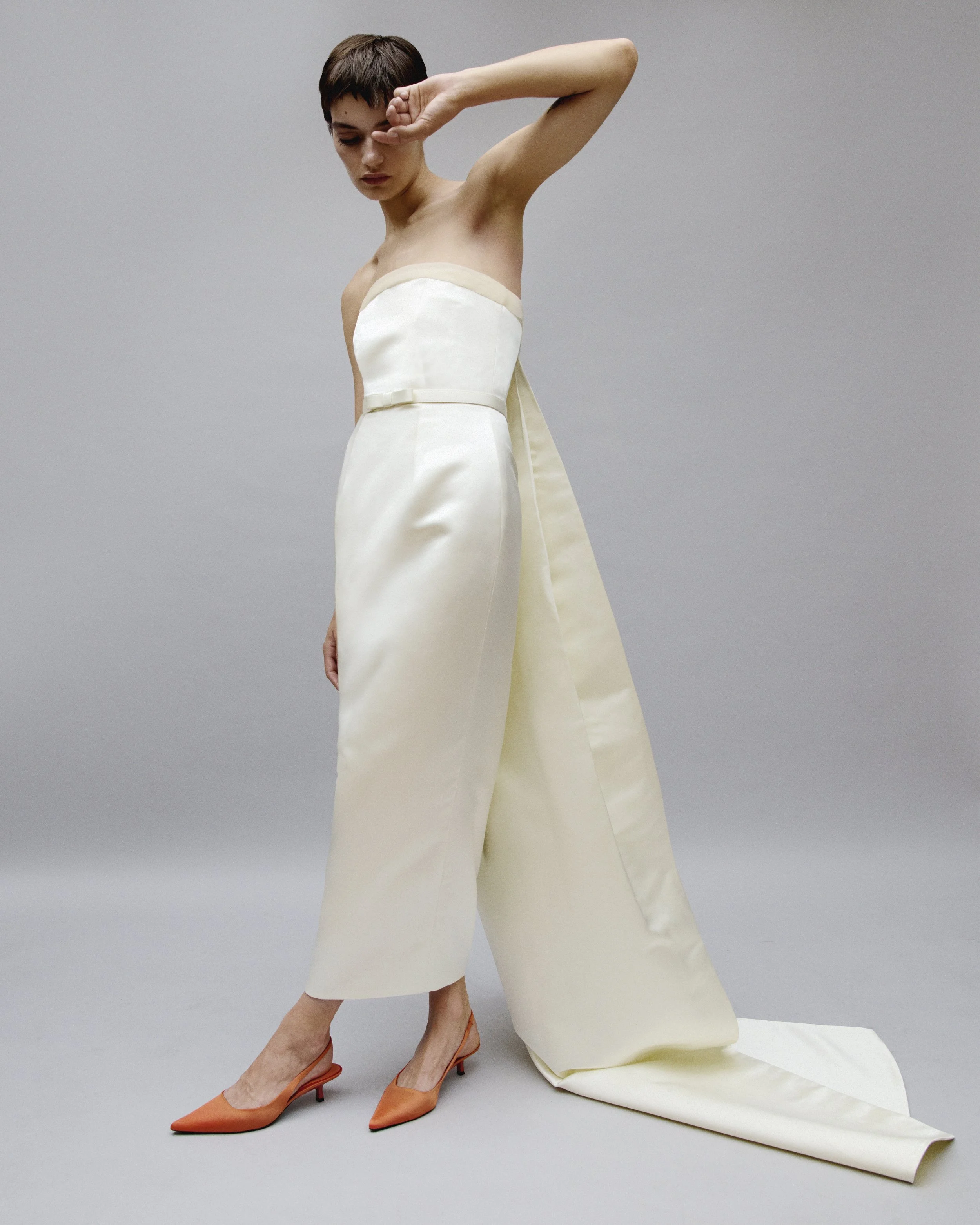 A woman in an off-shoulder white gown with a train, wearing orange high heels, standing against a gray background, with one arm raised and her hand resting on her forehead.