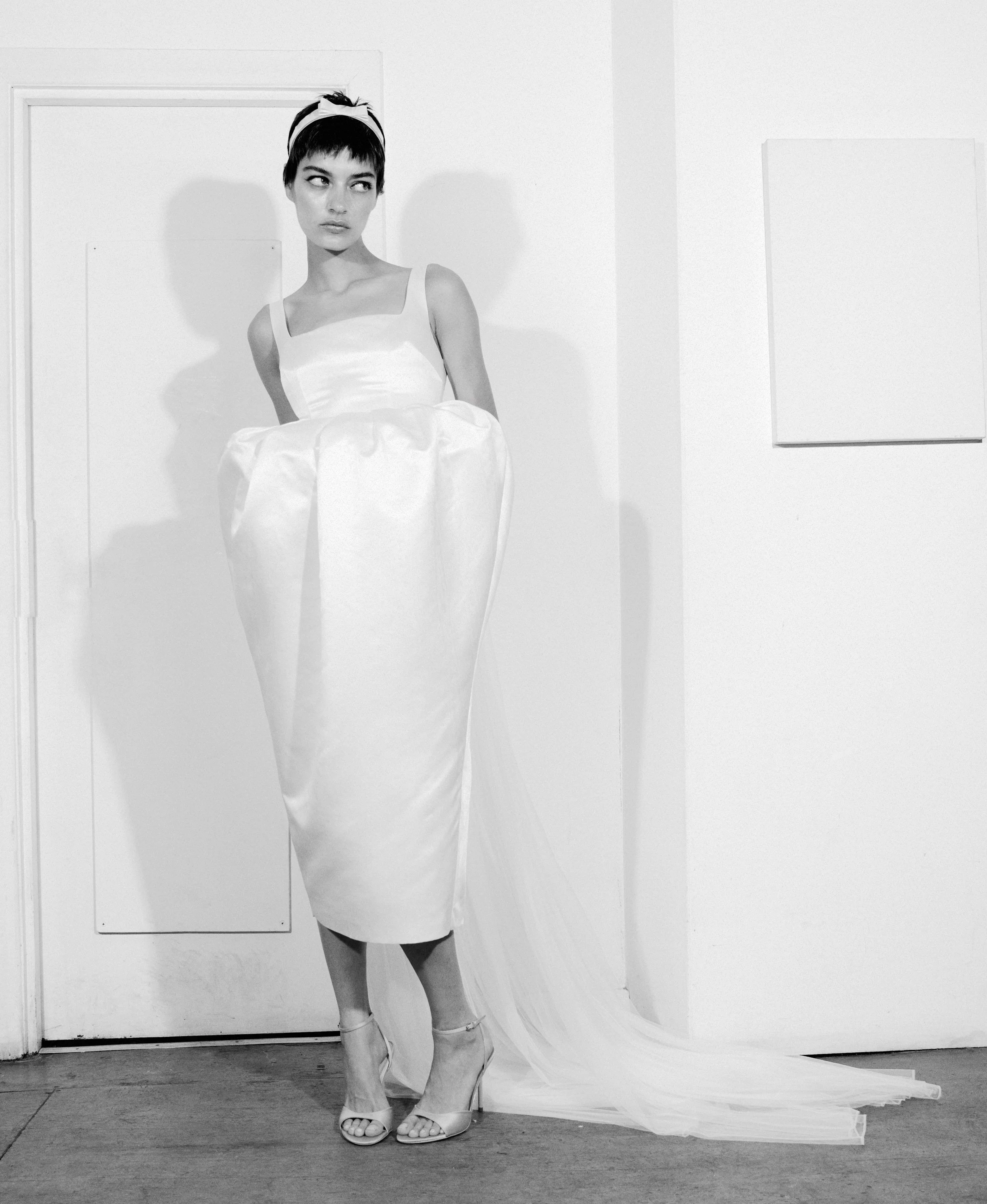Black and white photo of a woman in a white dress with a long train, wearing high heels and a headband, standing against a plain wall with shadows behind her.