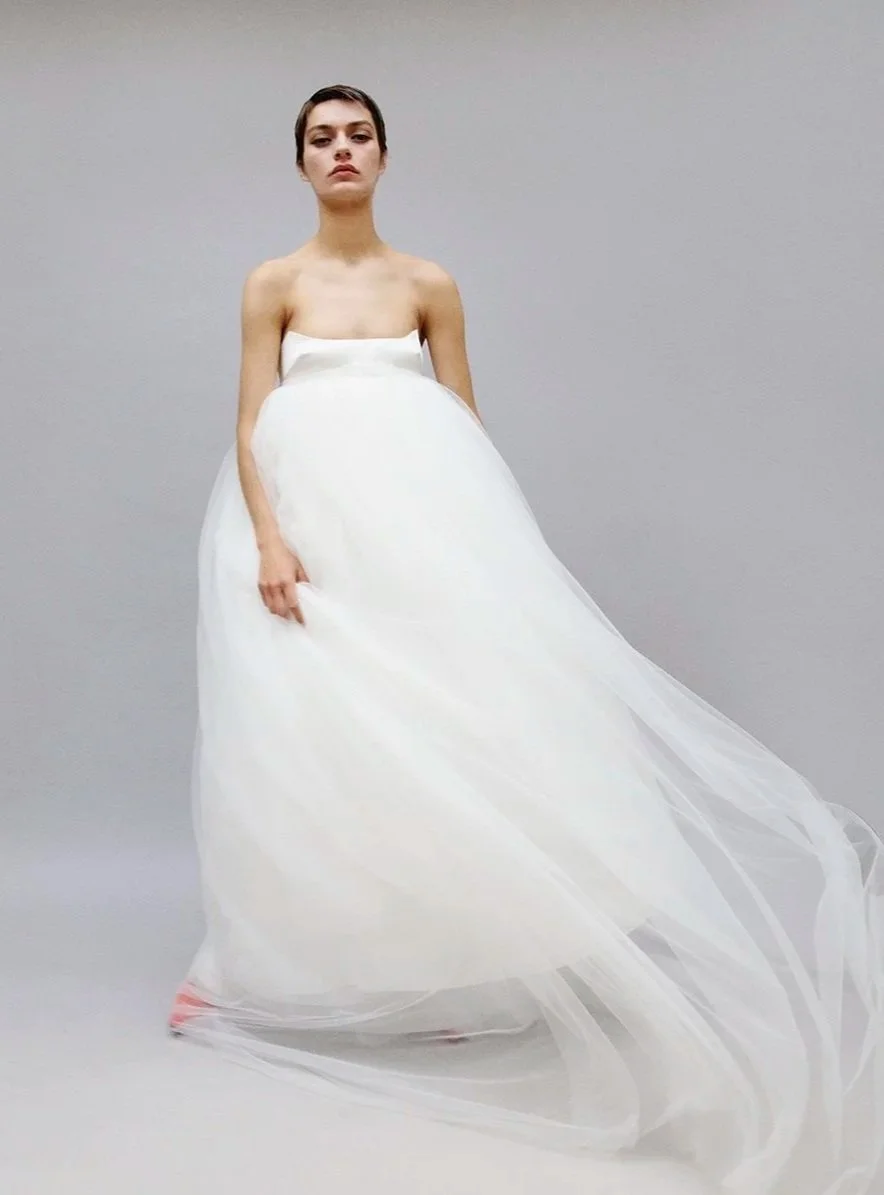 Model wearing a strapless white wedding dress with a full, flowing skirt, standing against a plain gray background.