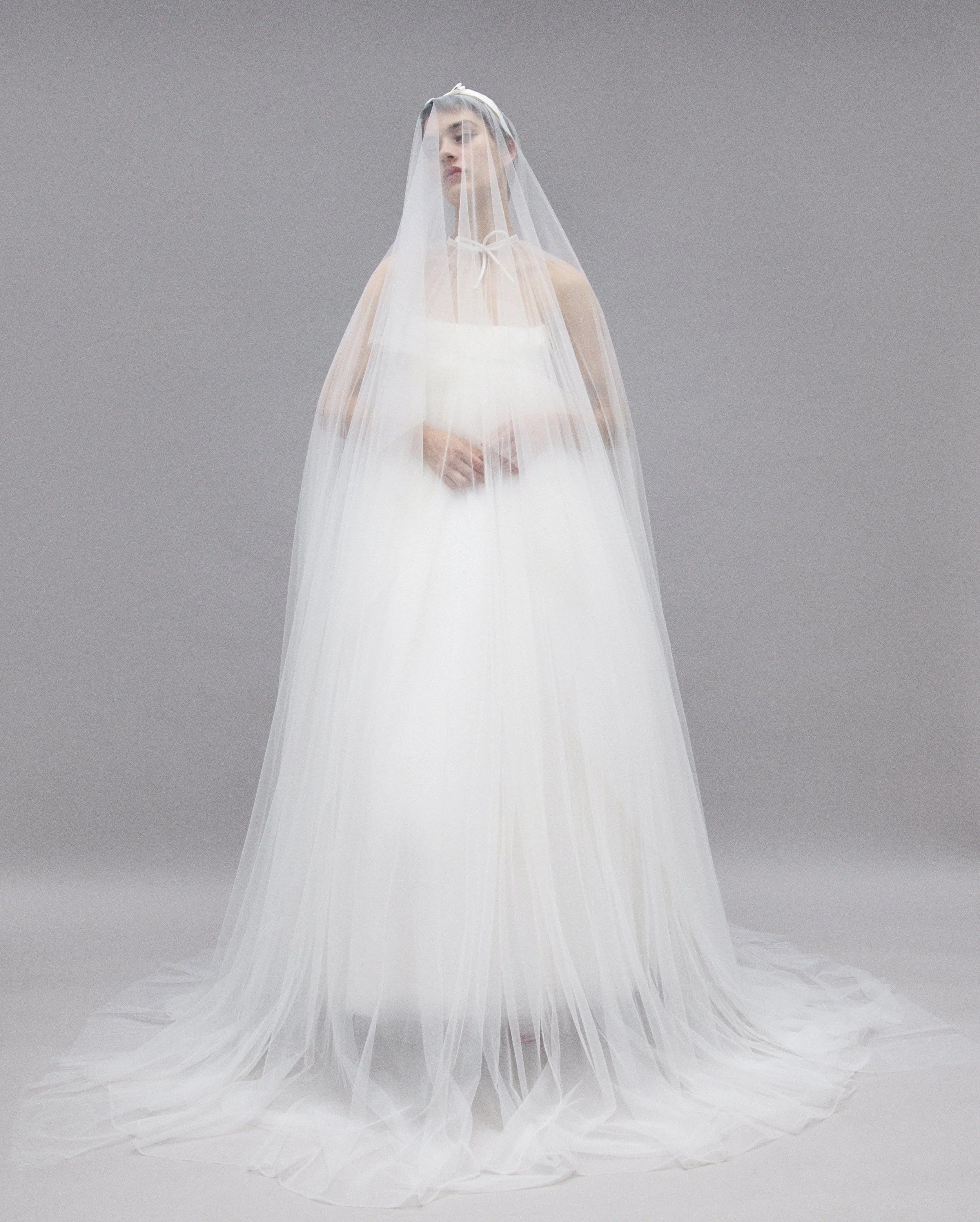 A woman standing in a white wedding gown with a sheer veil covering her face and upper body against a plain gray background.