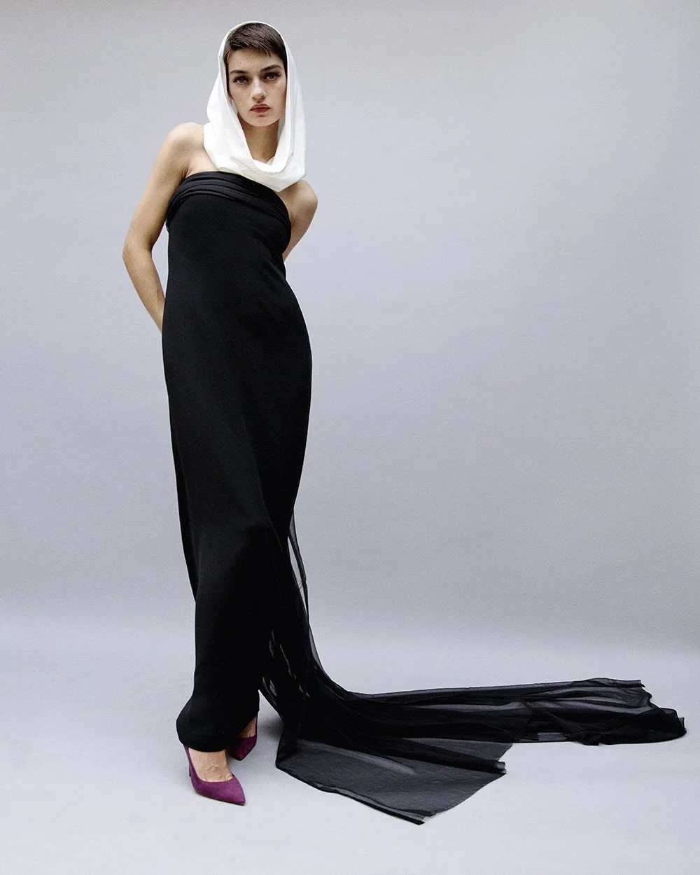 Fashionably dressed woman in a black strapless gown with a sheer train, purple heels, and a white headscarf, standing against a plain gray background.