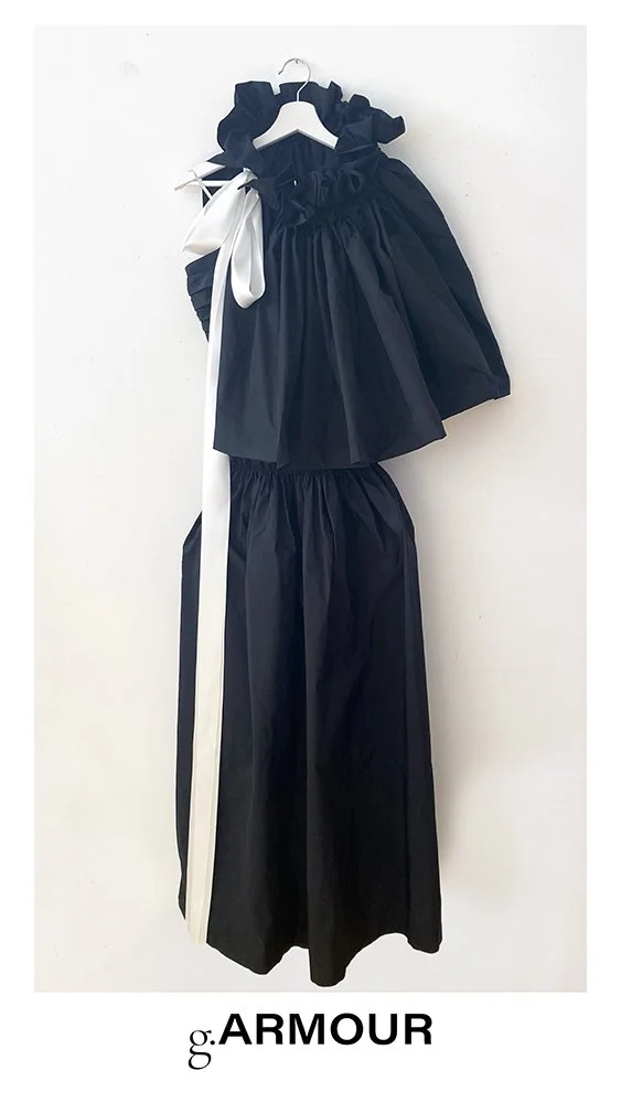 A navy blue dress and pants set hanging on a white hanger against a white wall. The dress has ruffled sleeves and a white bow on the shoulder. The pants are wide-legged with a white stripe down the side. The brand name 'g.ARMOUR' is at the bottom.