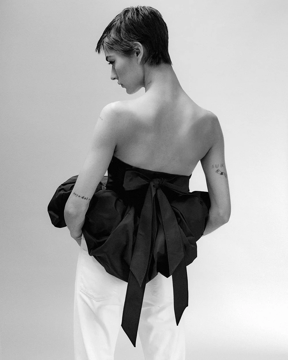 A woman with short hair looks down while holding a large black bag with a bow, wearing white pants, against a plain background.