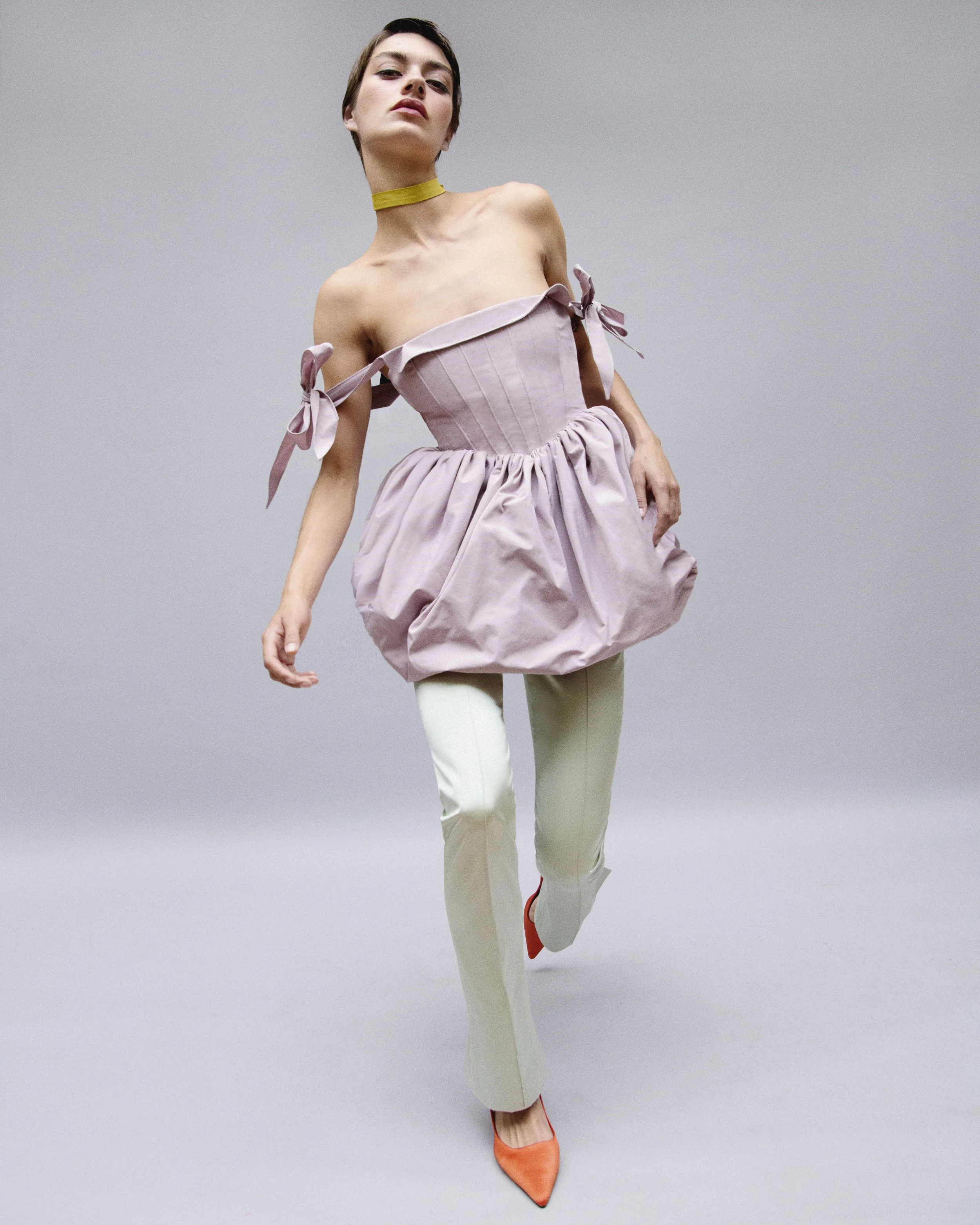 A woman in a pink off-shoulder dress with tied bows, cream pants, and orange heels posing against a gray background.