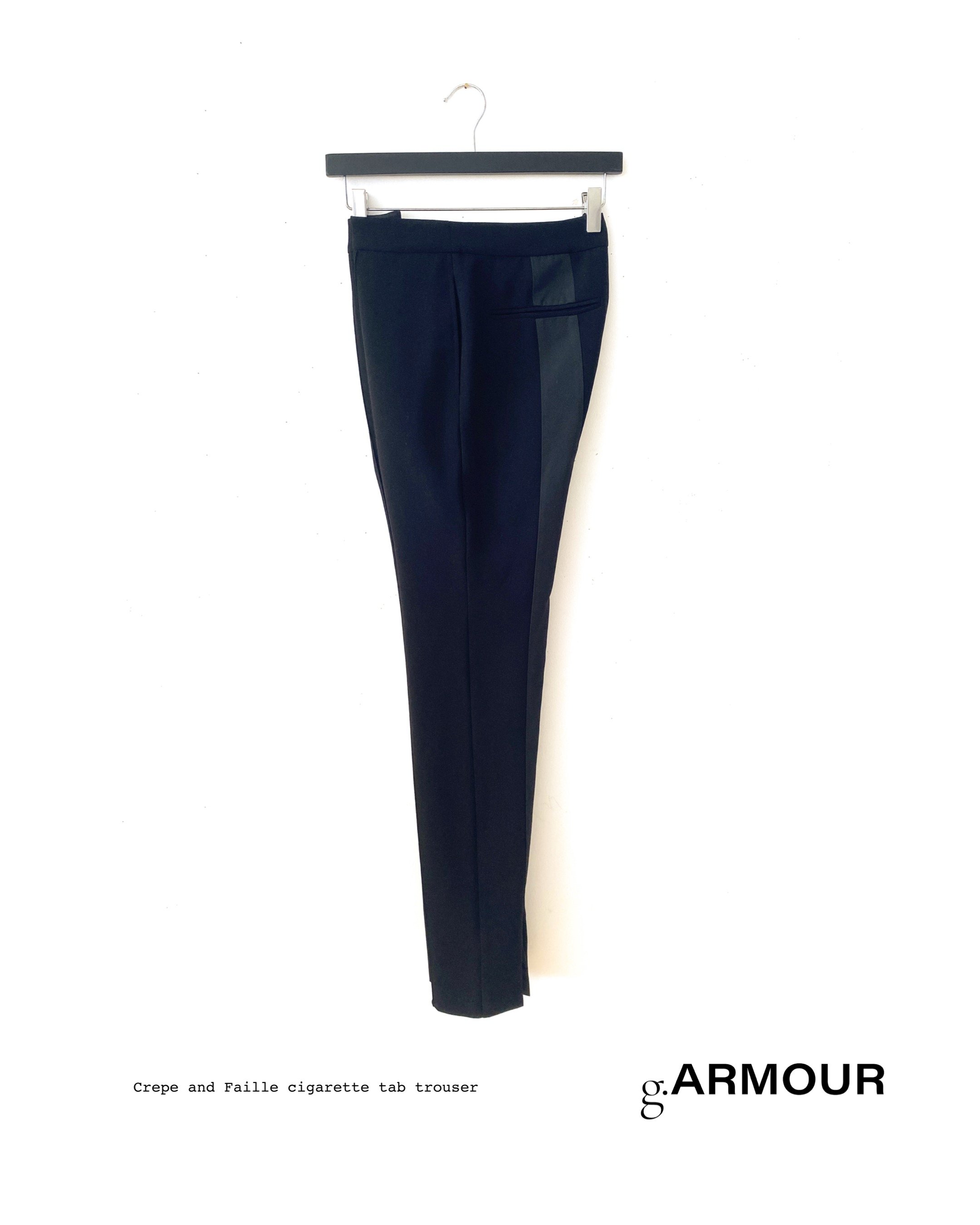 Black trousers with a leather stripe detail hanging on a black hanger, displayed against a white background with text indicating they are crepe and faille cigarette tab trouser from g.ARMOUR.