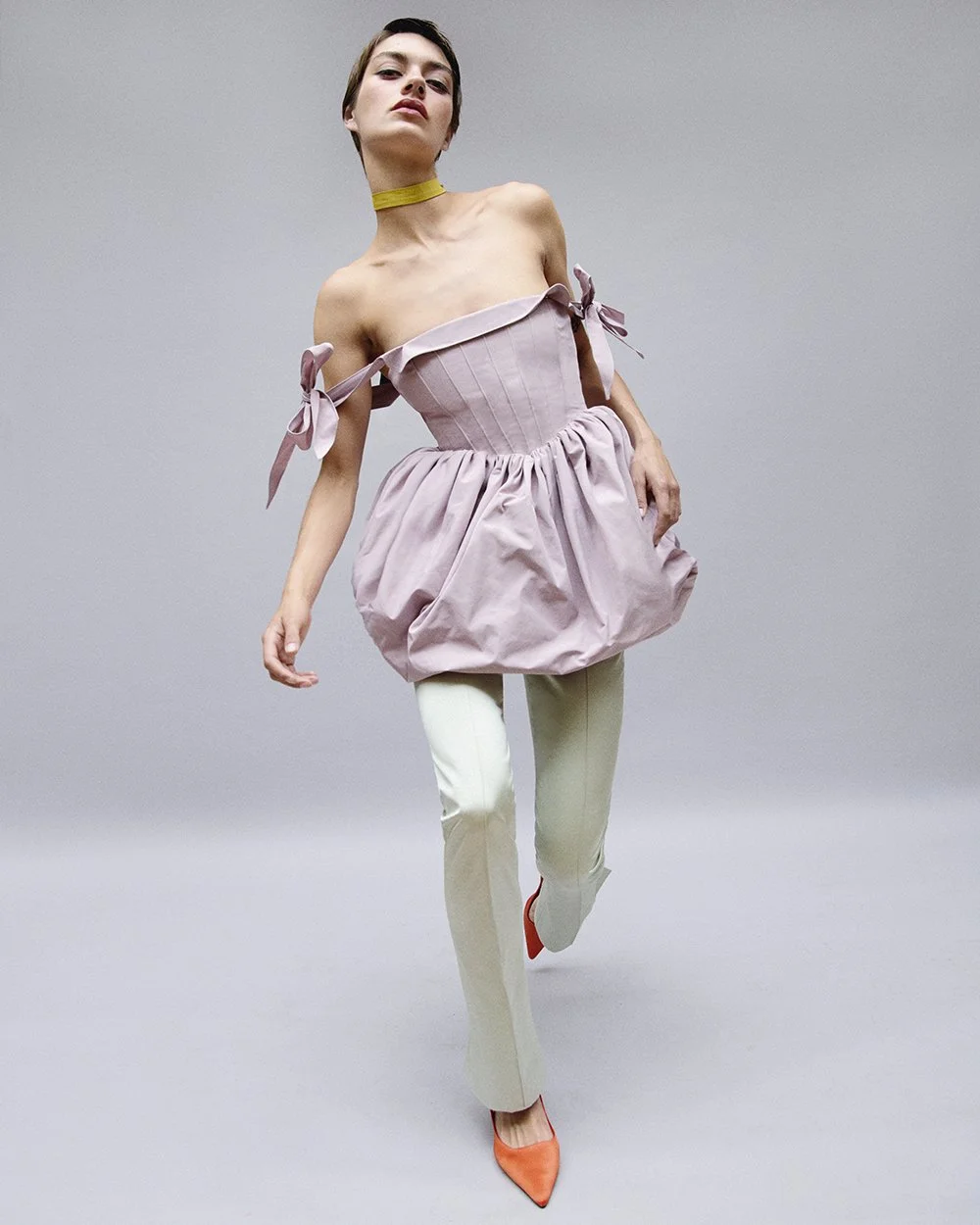 A woman with short hair dressed in a pink off-shoulder dress with ribbon bows, white pants, and orange shoes, standing against a plain gray background.
