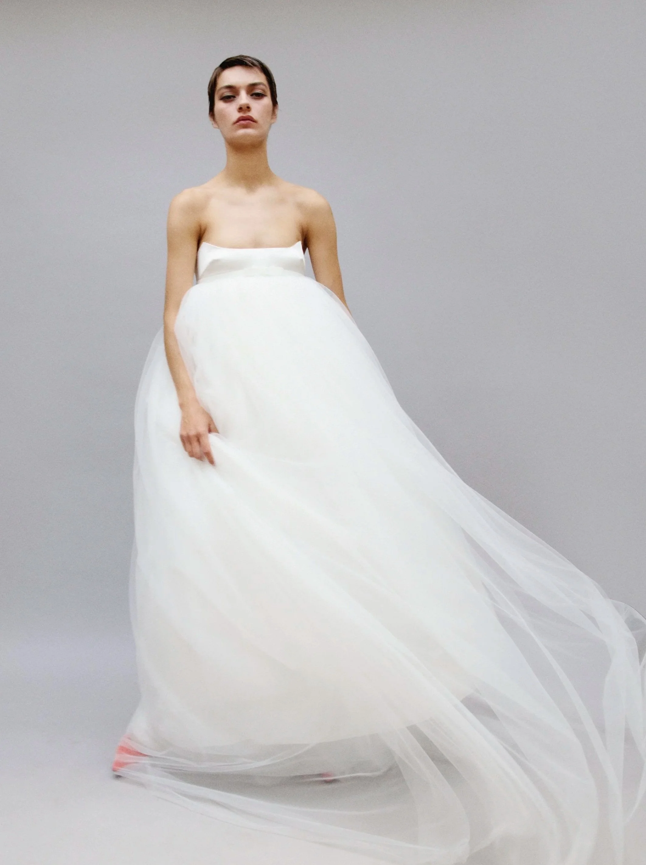 Young woman in a strapless white dress with a full skirt standing against a plain gray background.
