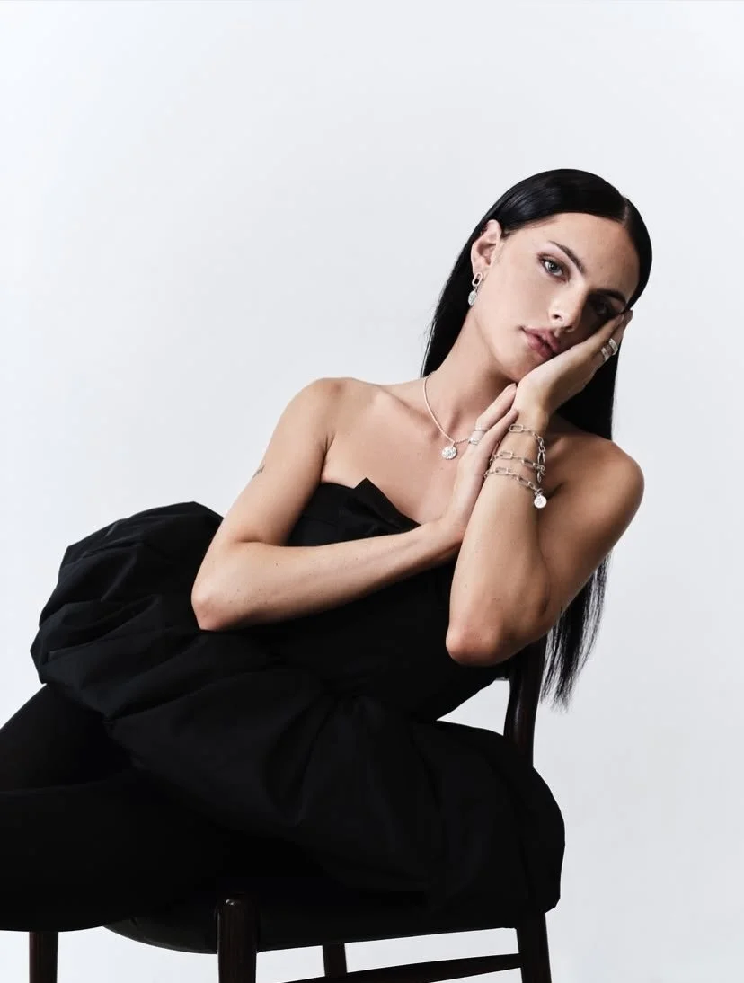 A young woman with long black hair, wearing a strapless black dress, sitting on a dark wooden chair with a white background. She is accessorized with jewelry including earrings, a necklace, and bracelets, and is resting her head on her hand.