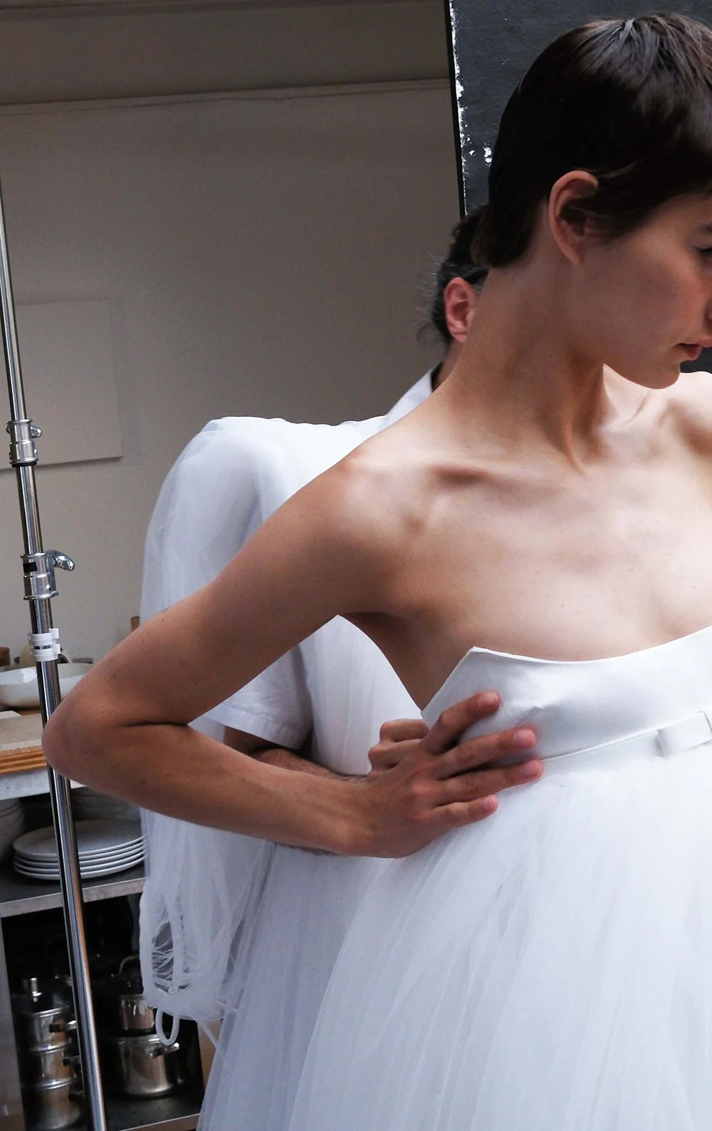 A woman is in a hospital gown, with her shirt slightly lifted, standing in a clinical setting, possibly a hospital. She has short brown hair and is looking downward, with a medical professional behind her adjusting her gown.