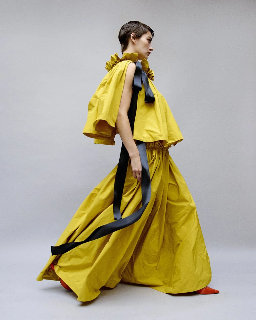 A fashion model in a yellow avant-garde outfit with oversized pants, a cape-like top, and black and red accessories, standing sideways against a neutral gray background.
