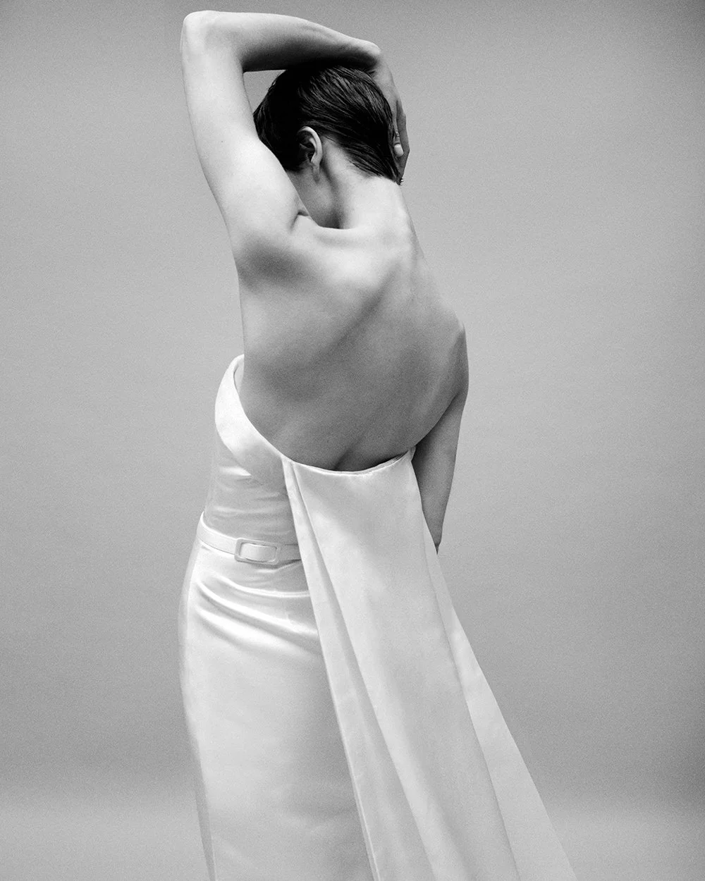 Black and white image of a woman with her back to the camera, her arms raised and hands resting on her head, wearing a white satin skirt.