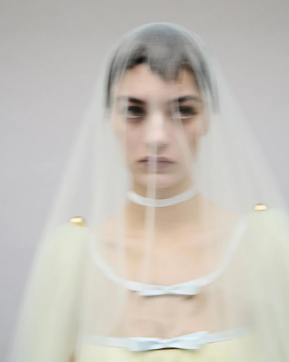 A woman with short dark hair, dressed in a light-colored top, is behind a transparent, slightly blurry veil or plastic sheet, creating a distorted and mysterious appearance.