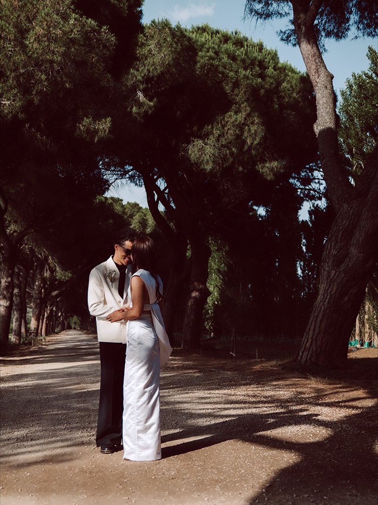 A couple dressed in white and black standing close together on a dirt path under large green trees.