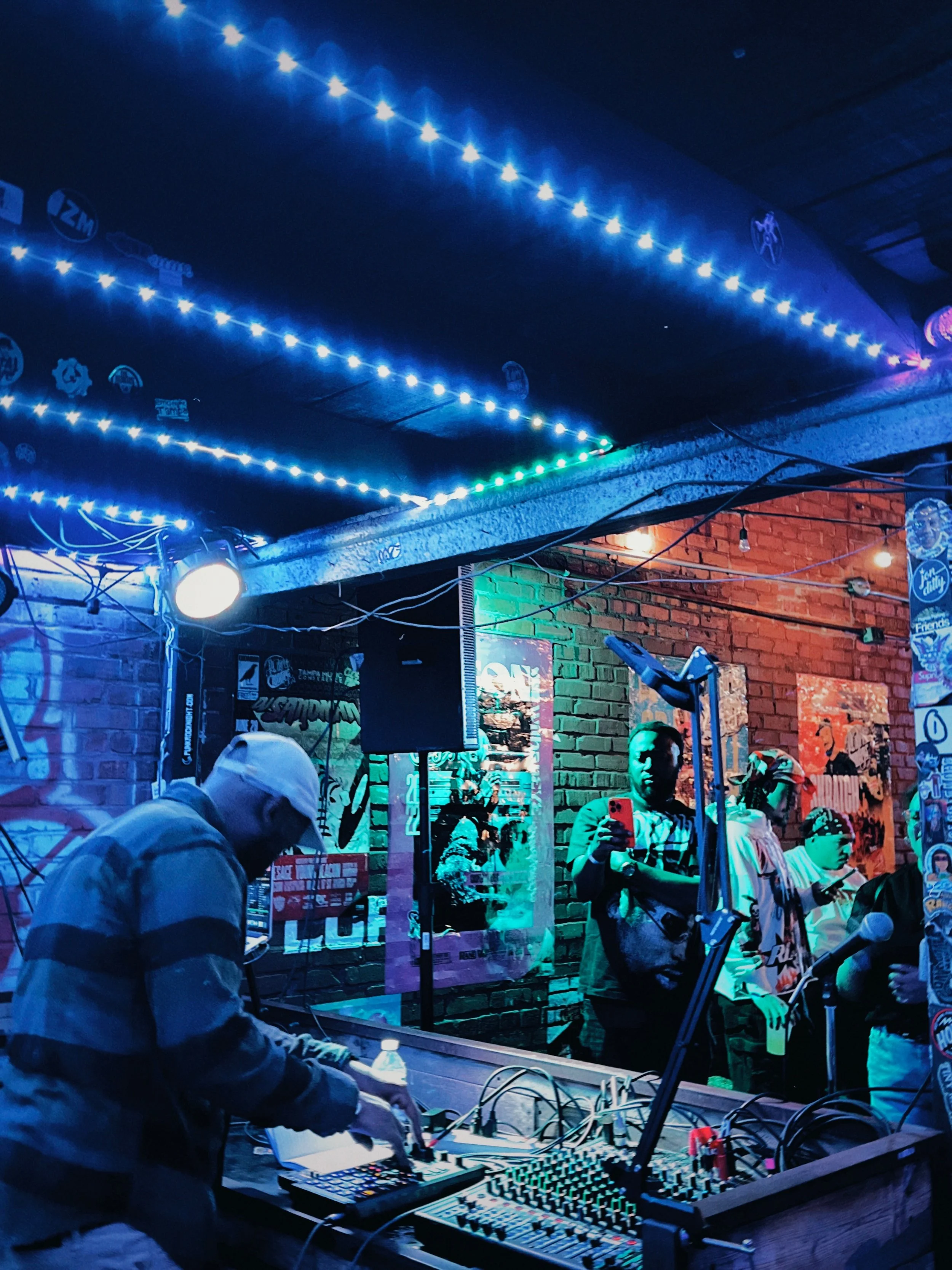 A DJ performs at an indoor music event with colorful LED lights, brick walls, and a small audience taking photos and listening.