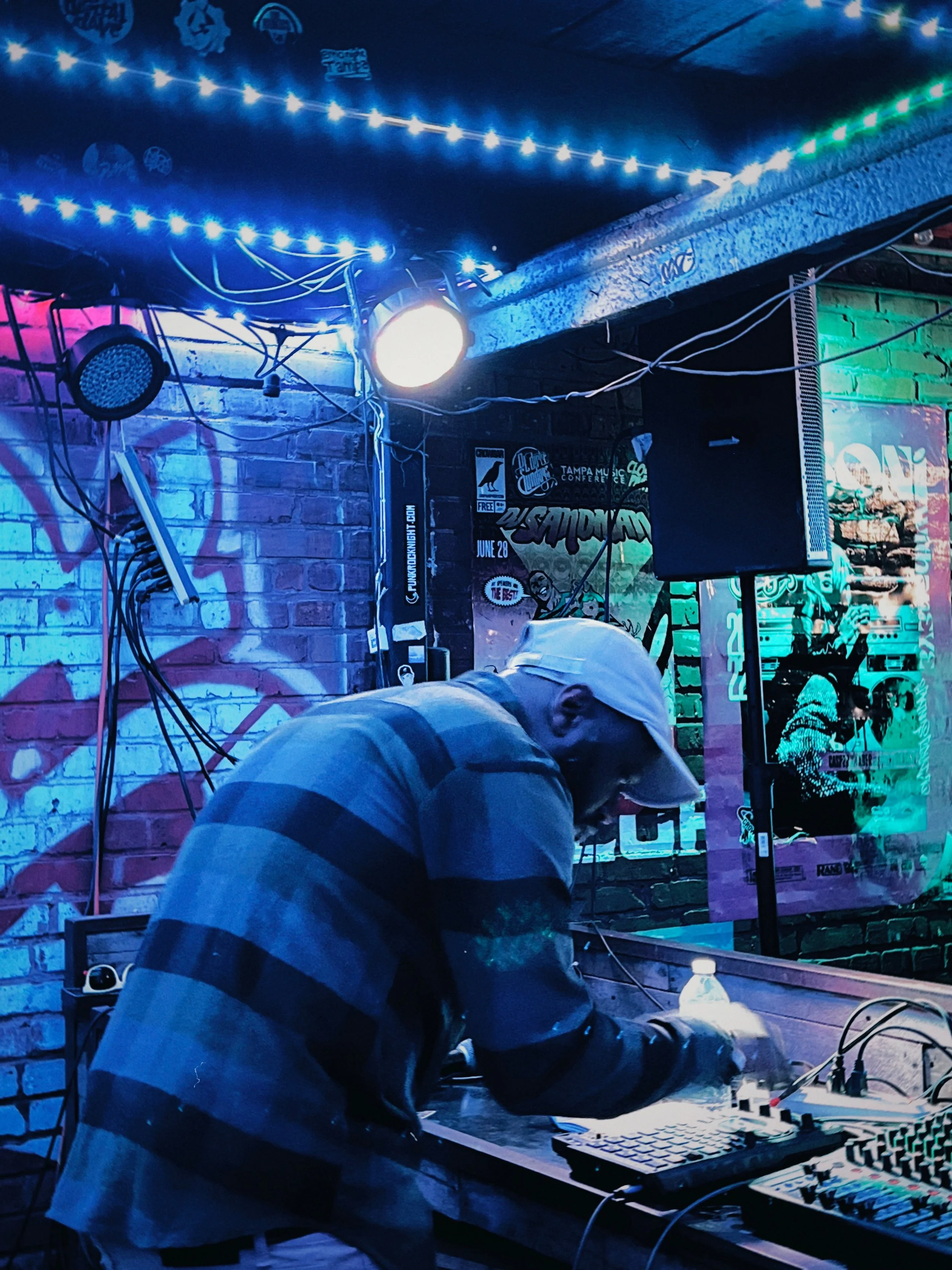 A DJ wearing a white cap and striped shirt performing at a venue with colorful neon and LED lighting, with posters on the brick wall behind him.