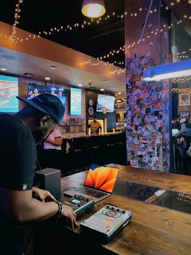 A DJ wearing a dark shirt and a cap performs with electronic music equipment at a bar or lounge decorated with string lights, posters, and a collage of stickers on a pillar. Several TVs are mounted on the wall displaying advertisements for billiards,
