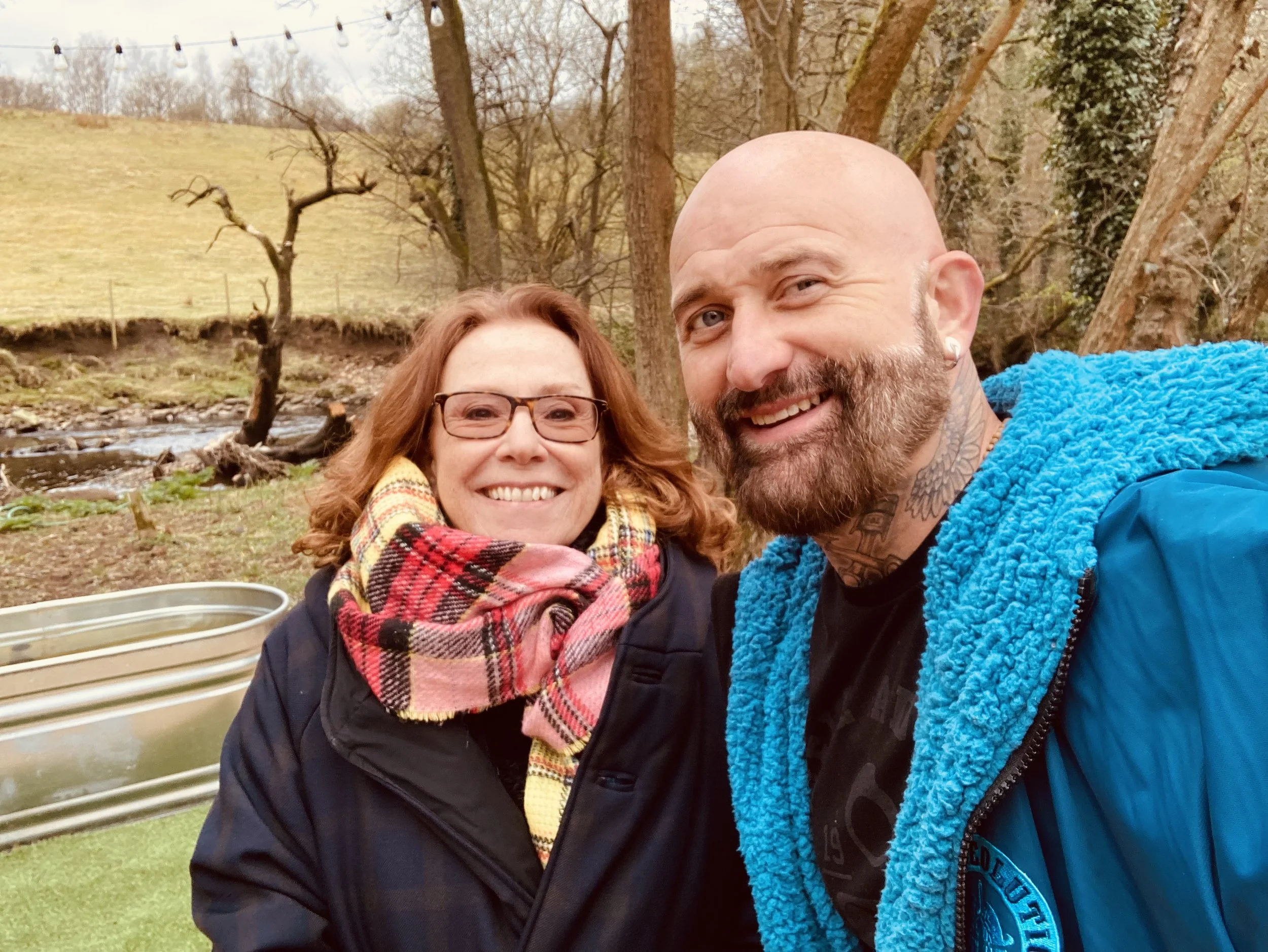A smiling woman with glasses and colorful scarf, and a smiling man with beard and tattoos, taking a selfie outdoors near a small stream in a grassy, wooded area.