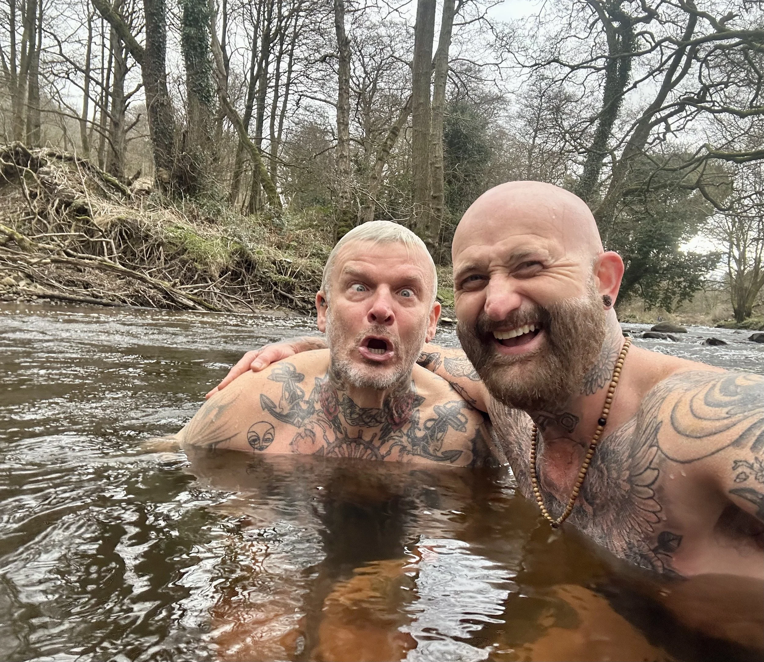 Two tattooed men with facial hair are shirtless and in a river, making funny faces at the camera. They are outdoors with trees and a rocky bank in the background.