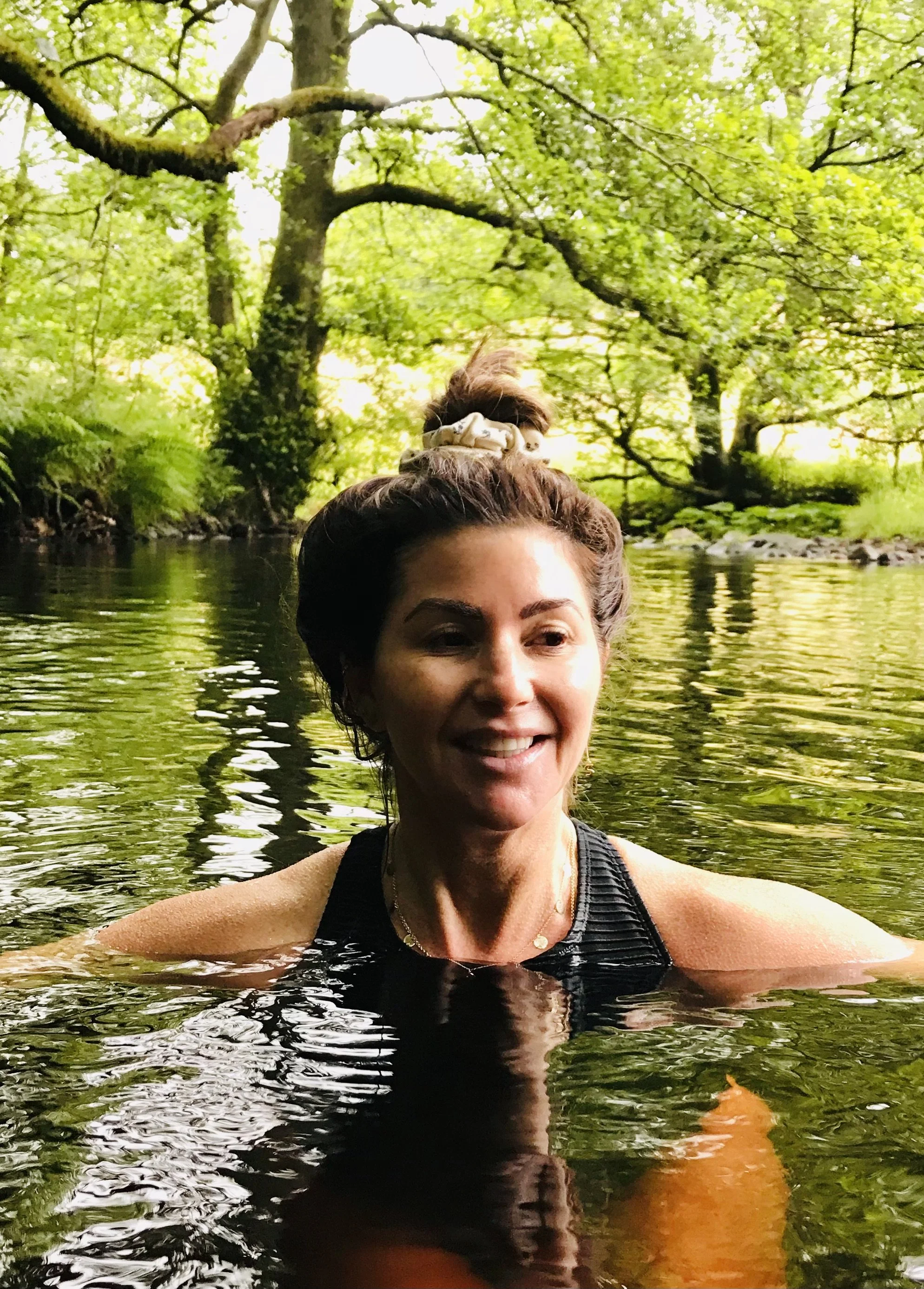 A woman with dark hair tied in a bun, smiling while swimming in a river surrounded by lush green trees.