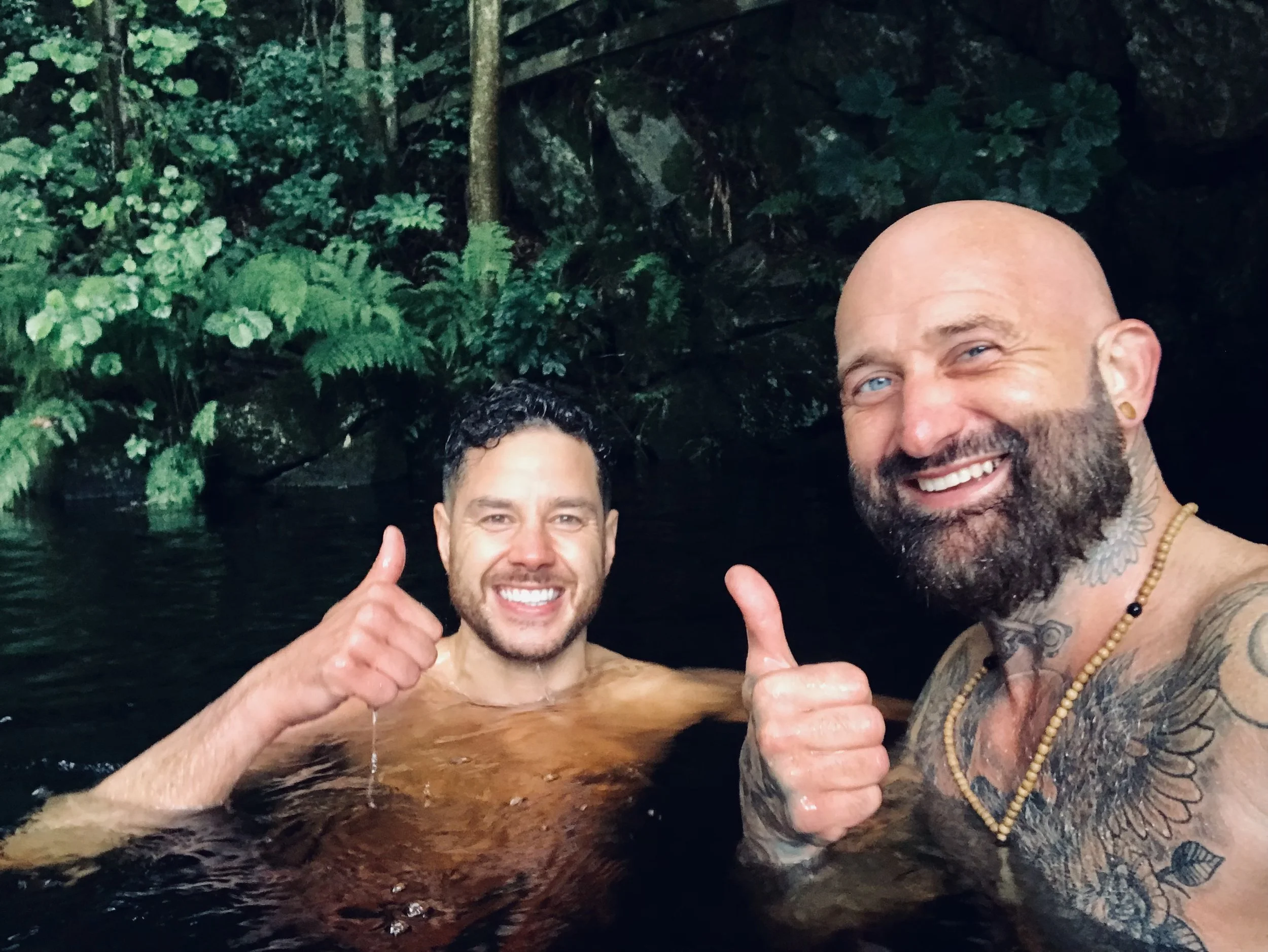 Two men smiling and giving a thumbs-up while partially submerged in a natural pool or hot spring, surrounded by lush greenery and rocks.