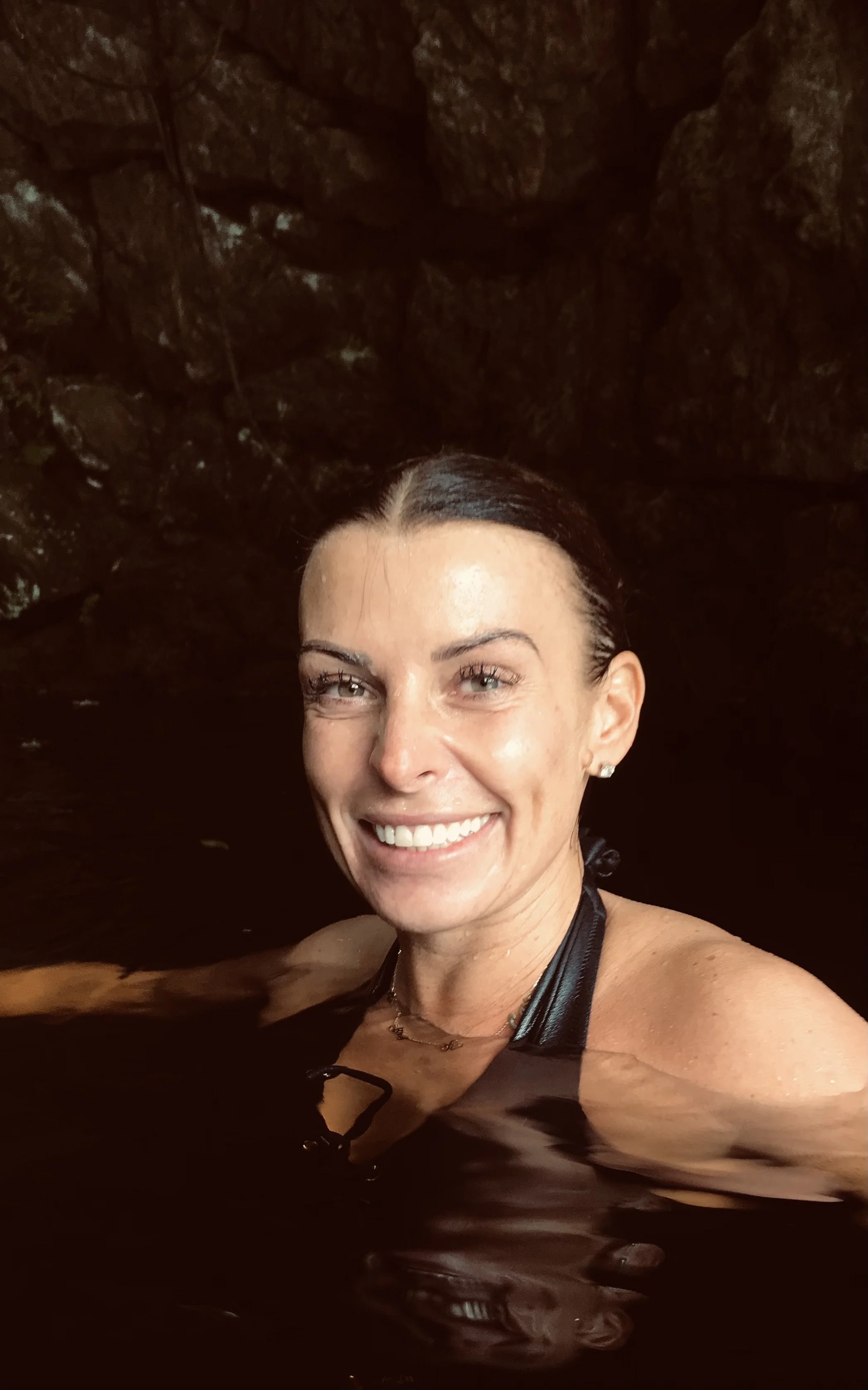 A woman smiling while swimming in a dark water body with rocky surroundings.