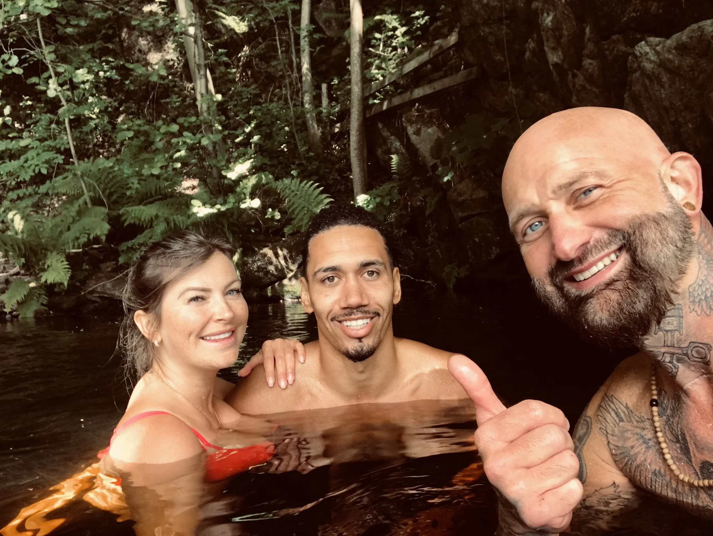 Three people, two men and a woman, smiling and taking a selfie together while swimming in a natural water body surrounded by green foliage and trees.