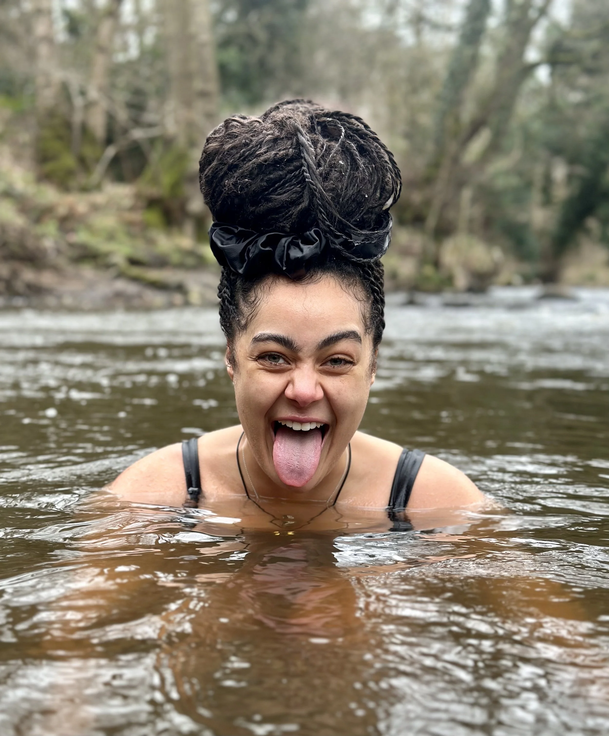 Woman with braided hair in a bun, wearing a black hair scrunchie, sticking out her tongue and smiling while standing in a river during daytime.