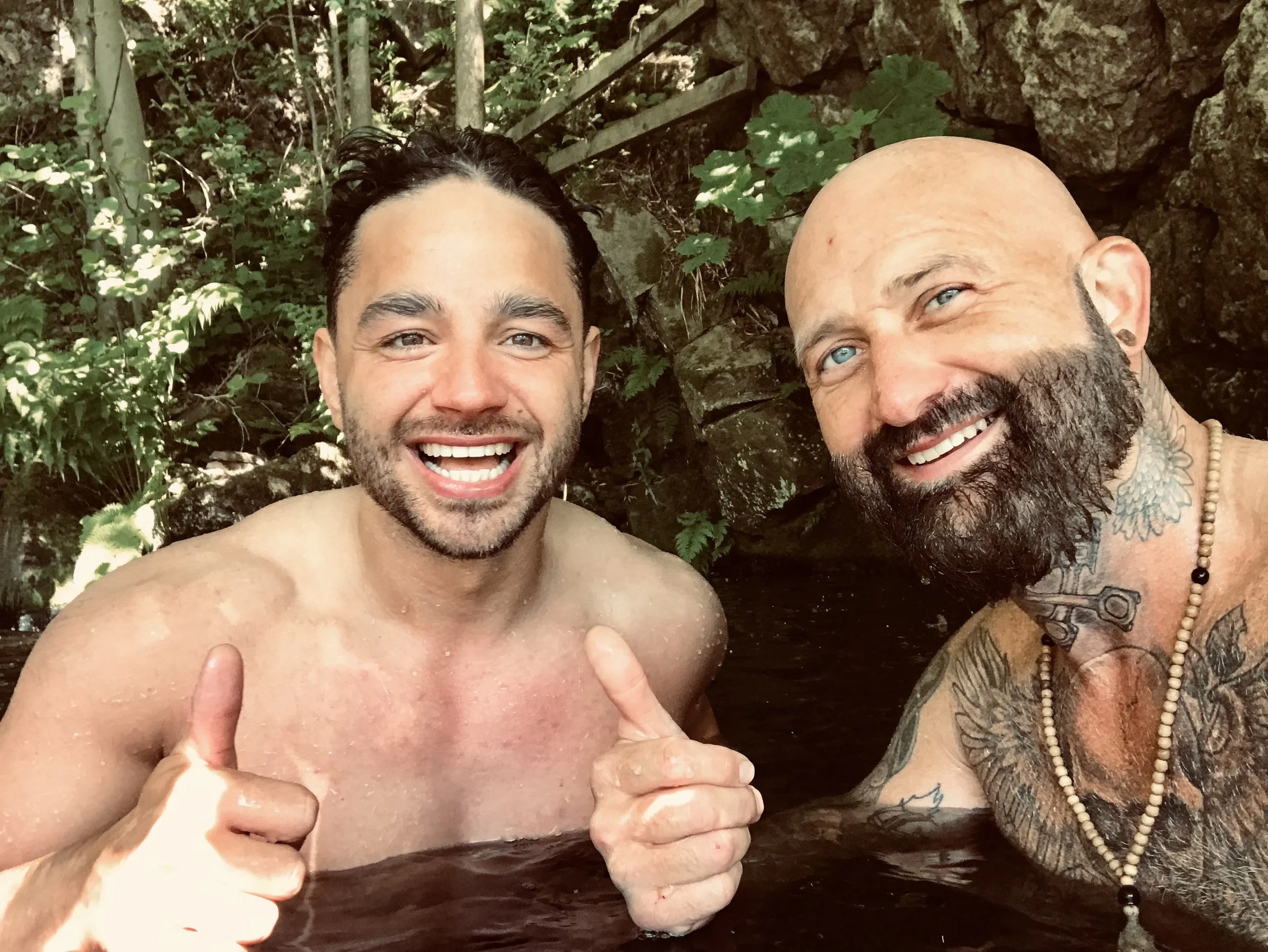 Two smiling men in a natural outdoor setting, partially submerged in water. One is giving a thumbs-up. They are surrounded by greenery and rocks.