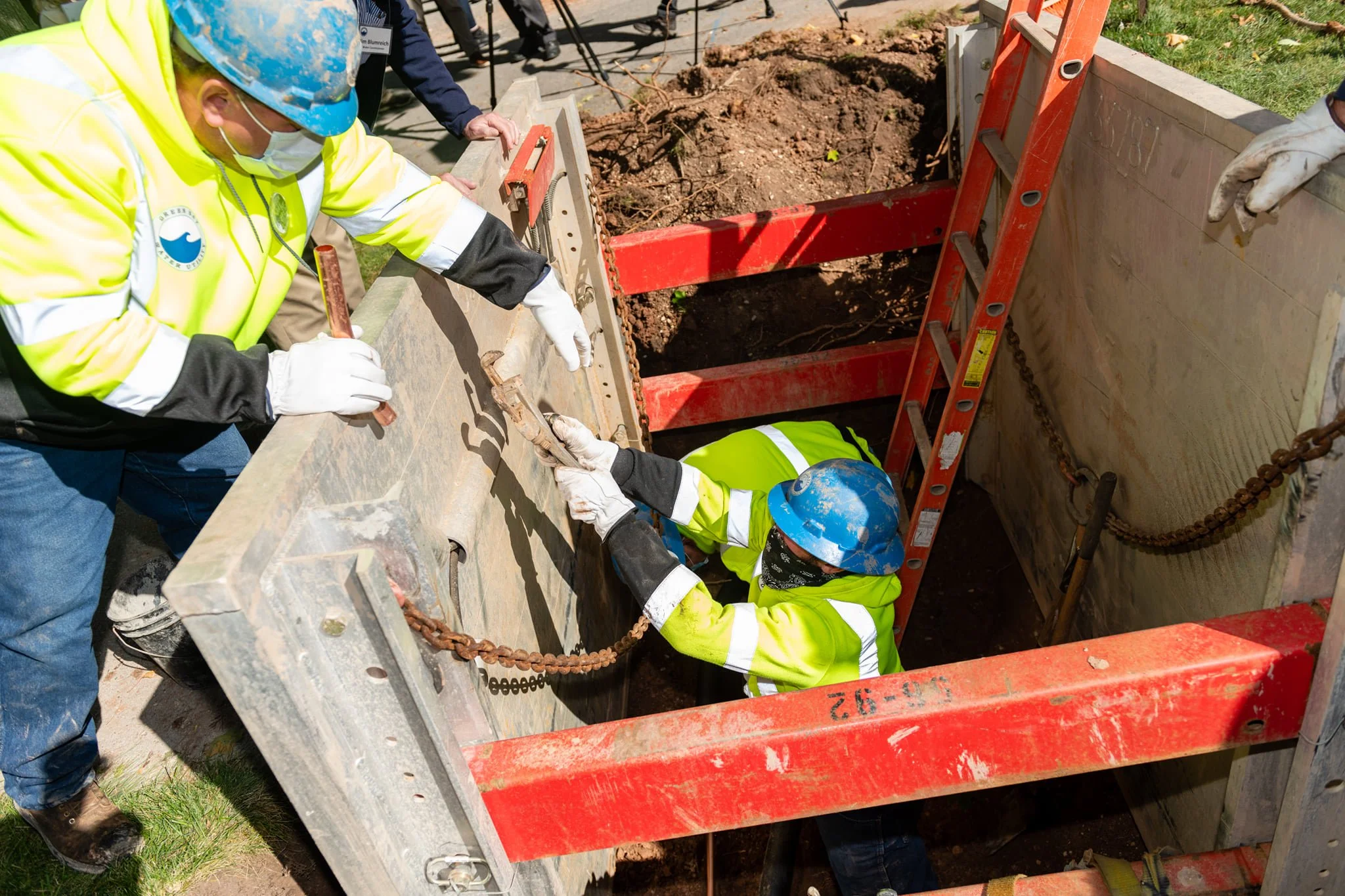 Two construction workers in yellow safety jackets and blue helmets are installing a large concrete structure into the ground, with one worker inside the trench and the other outside, both securing the structure.