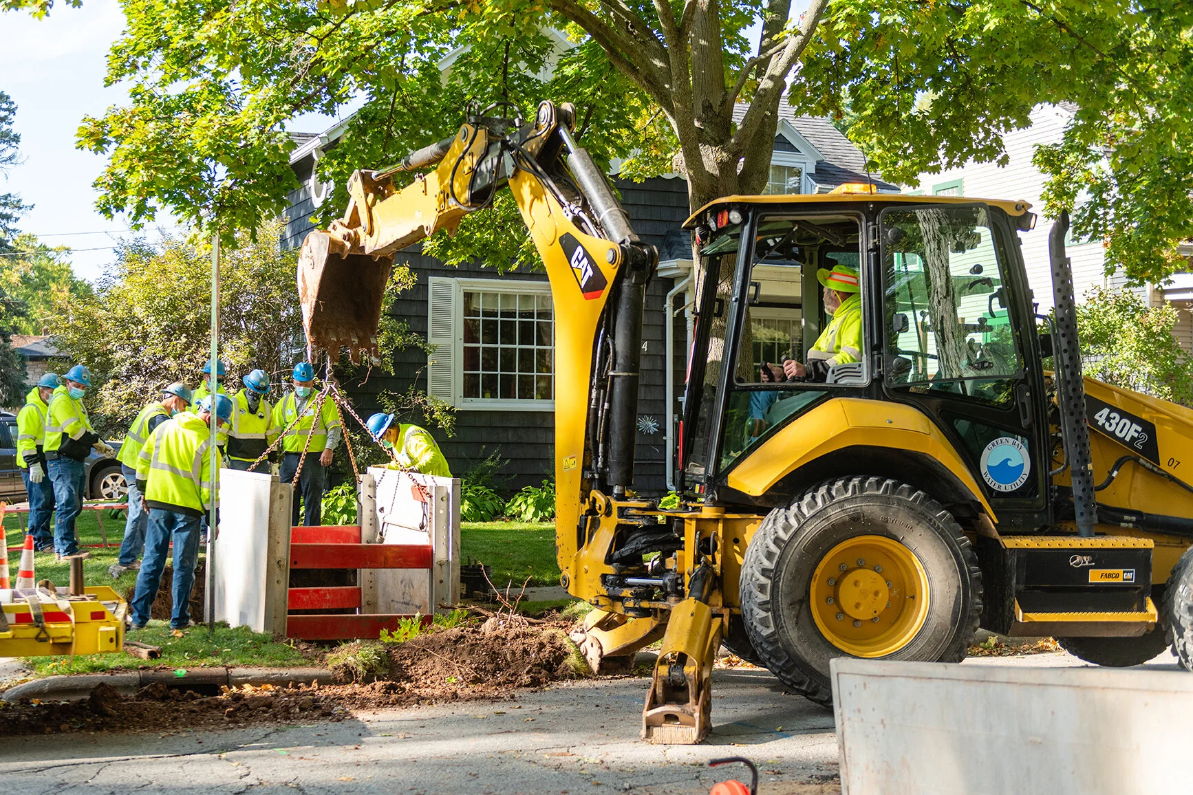 Construction workers in yellow safety vests and helmets operate a backhoe to dig a trench in a residential area, with a black house and large trees in the background.