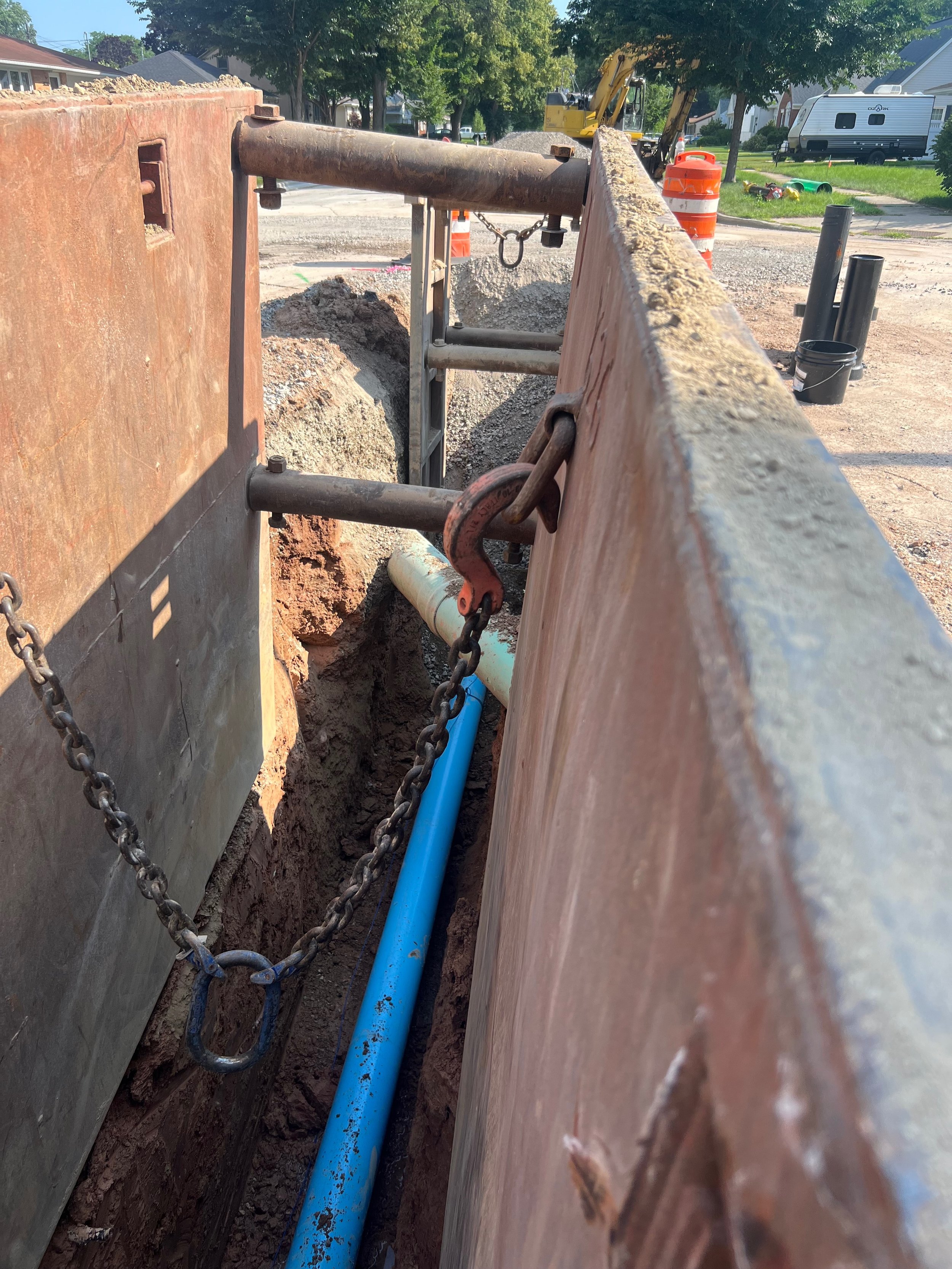 Construction site with dirt trench showing a blue underground pipe and construction barriers, with trees and houses in the background.