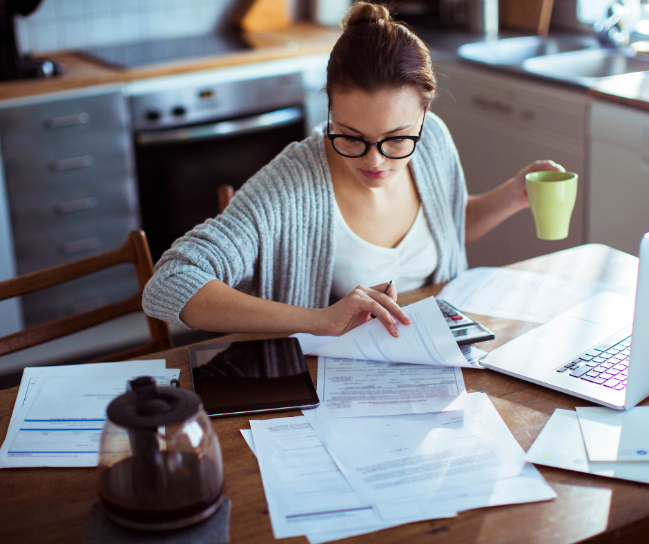 A woman with glasses sitting at a kitchen table working on financial documents, holding a yellow coffee mug in her left hand, surrounded by papers, a tablet, a laptop, and a calculator.