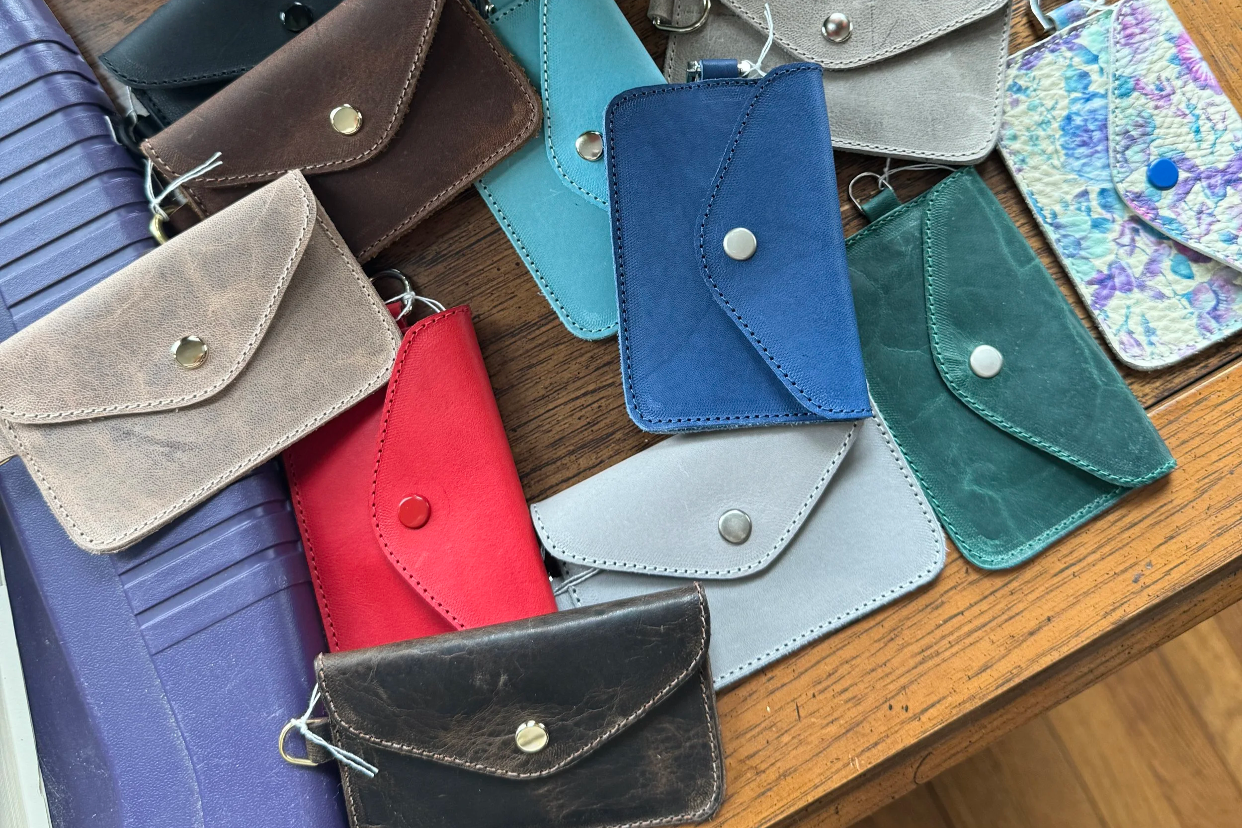 A collection of colorful wallets and cardholders spread out on a wooden surface. The wallets are in various colors including purple, beige, red, brown, blue, green, and metallic. Some wallets are textured, and they have snap buttons or clips.
