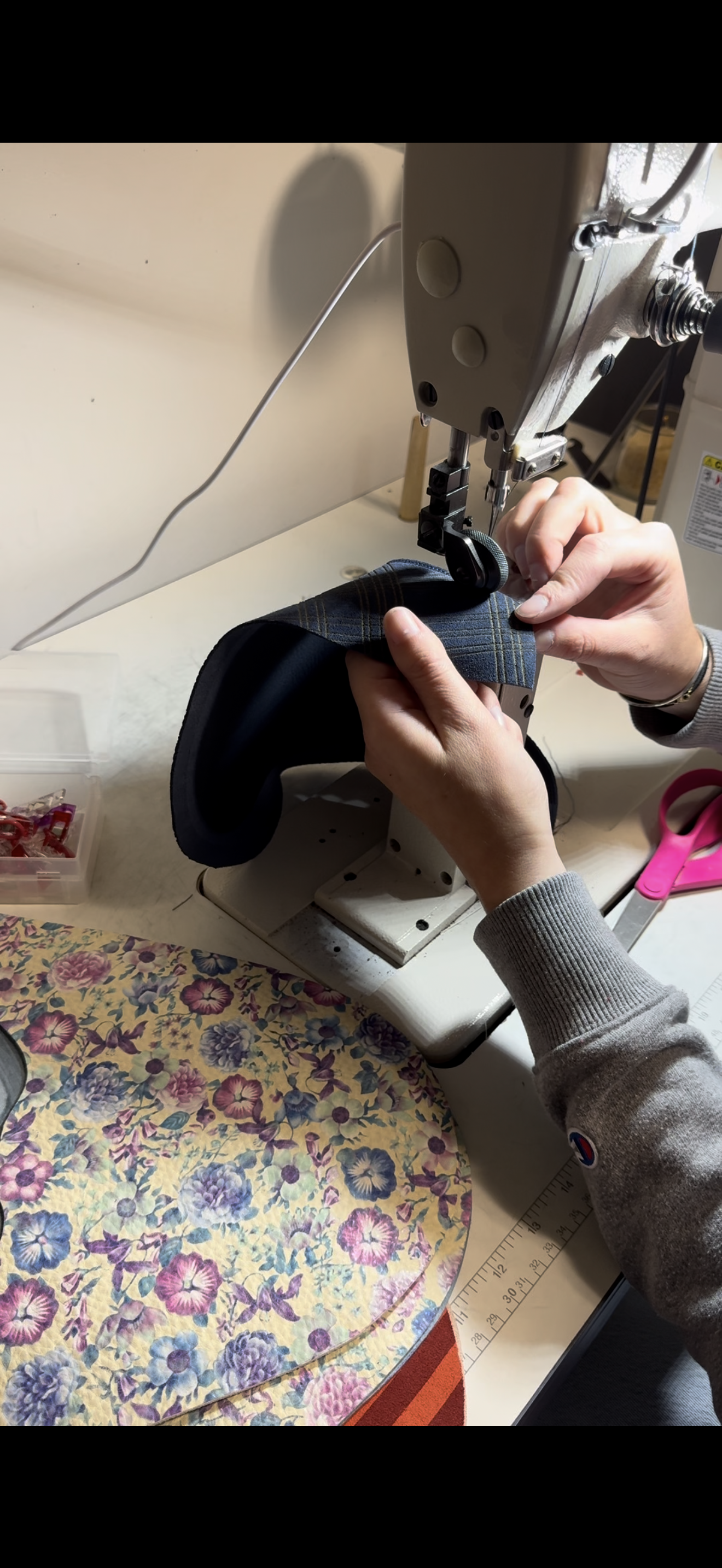 Person sewing fabric with a sewing machine on a worktable. There is a floral fabric pattern and sewing supplies around.