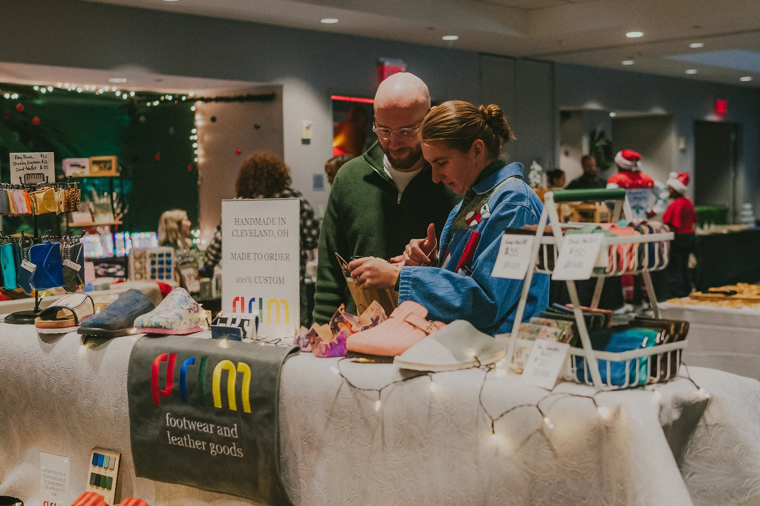 Two people browsing at a holiday-themed gift market stall decorated with Christmas lights and ornaments. The stall features footwear, leather goods, and other handmade items, with signs indicating custom orders and pricing.