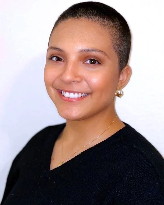A woman with a shaved head, wearing gold earrings and a black top, smiling against a plain white background.