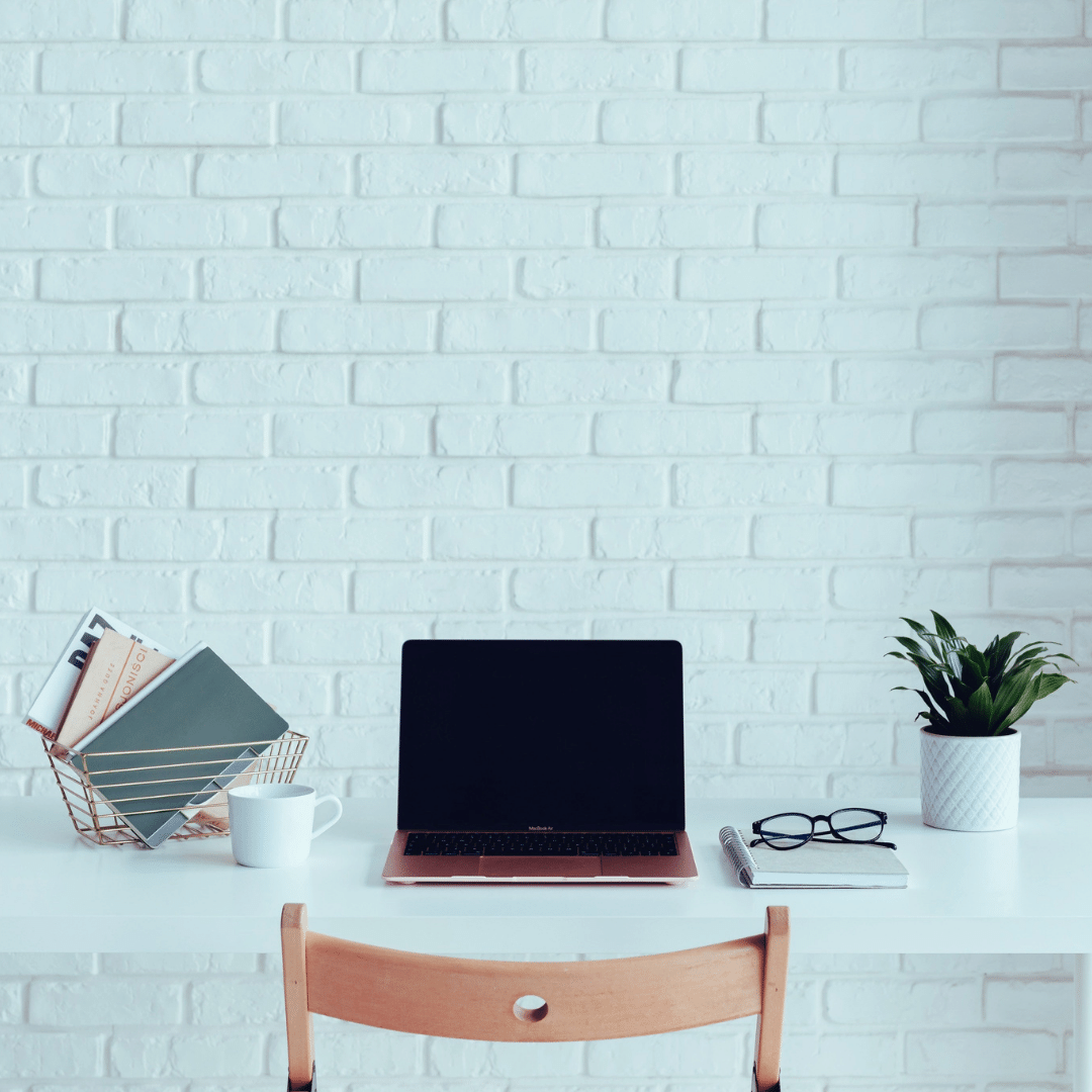 Minimalist workspace with laptop, books, plant, and coffee mug on white desk against a white brick wall.