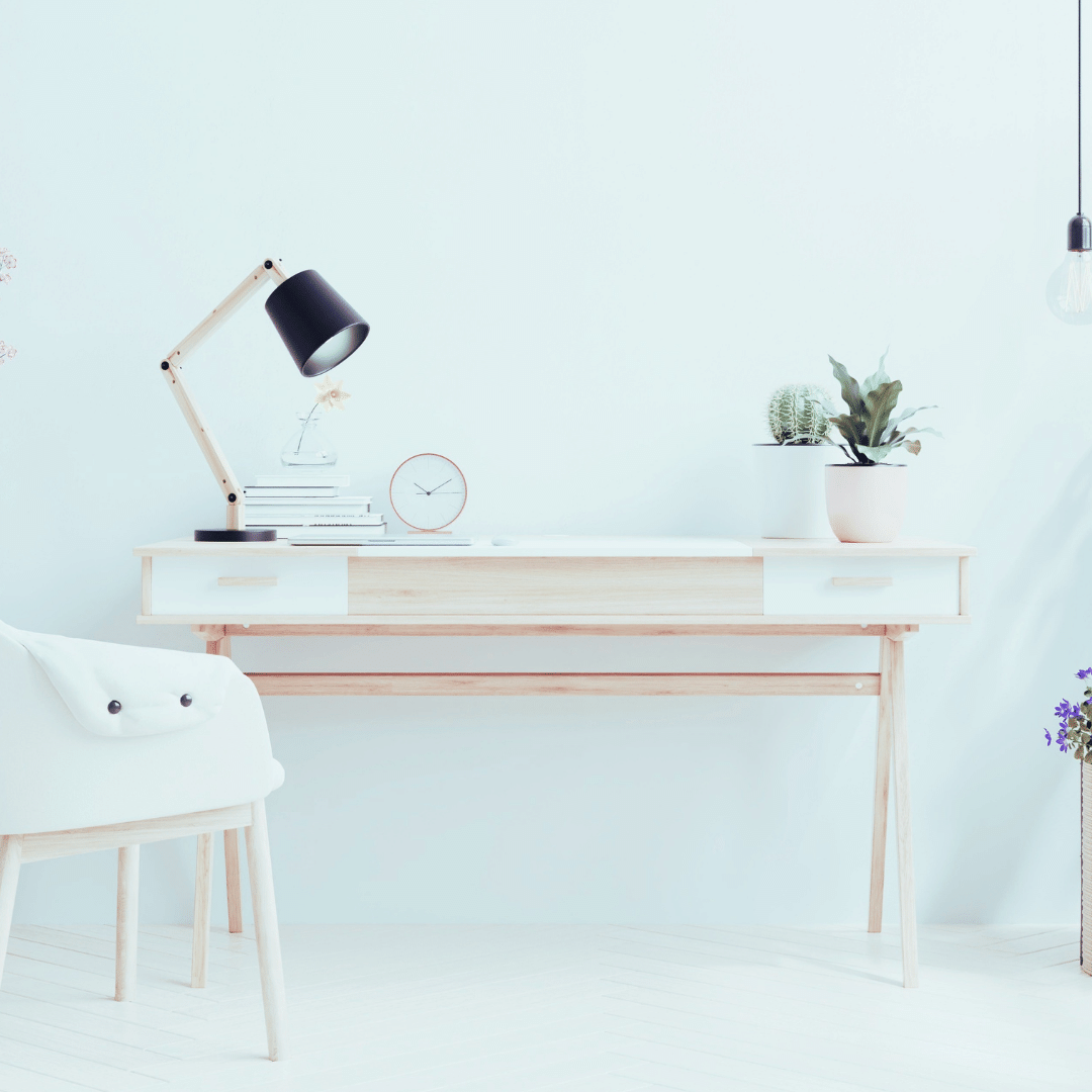 Minimalist workspace with a wooden desk, white chair, desk lamp, clock, books, and two potted plants in a light-filled room.