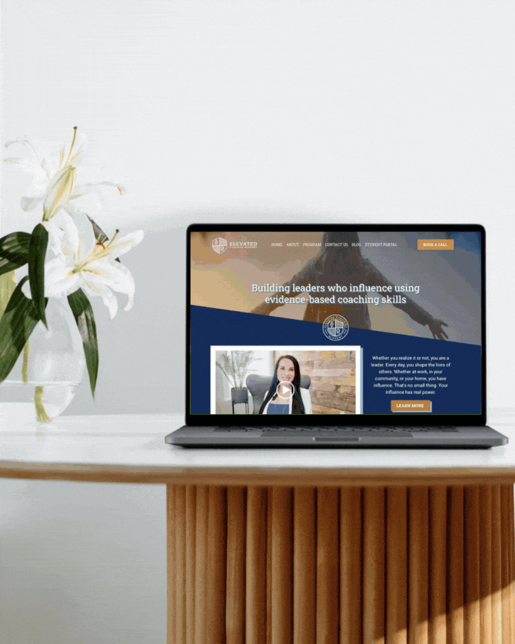 Laptop on a wooden table displaying a coaching website, with a vase of white lilies beside it.