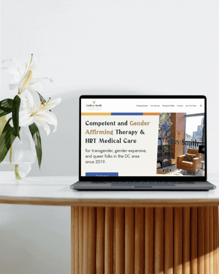 Laptop displaying a website for counseling and gender-affirming therapy, placed on a wooden table next to a vase of white lilies.