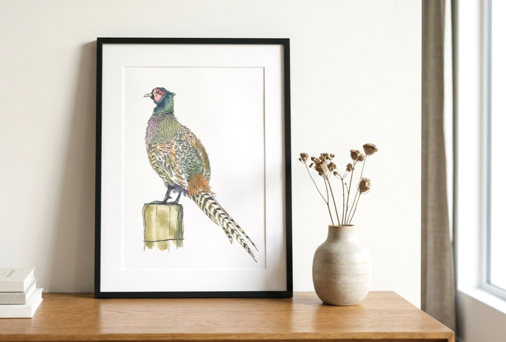 A framed illustration of a pheasant on a wooden surface, next to a beige vase with dried flowers, in a bright room with a window and curtains.