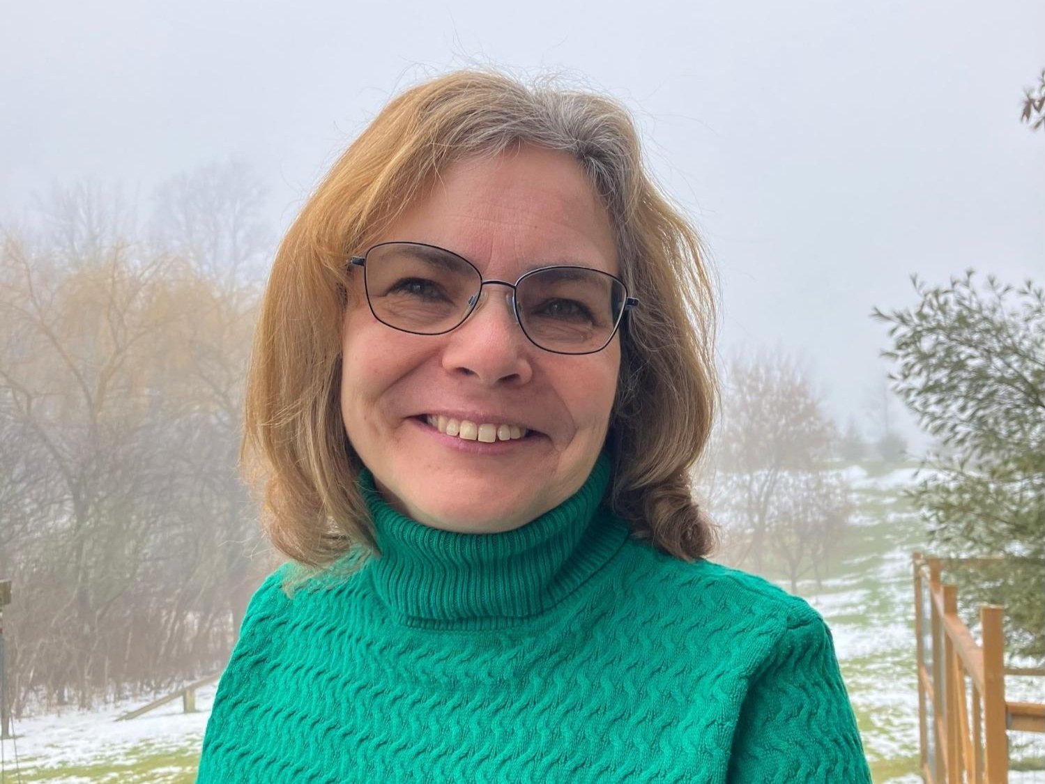 Woman wearing glasses and green sweater smiling outdoors in a foggy landscape.