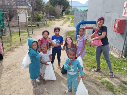 Group of smiling children and one adult holding bags on a dirt path next to a building, with trees and a field in the background.