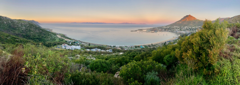 Panoramic view of a coastal landscape with lush greenery, a bay, and mountains in the background under a clear sky during sunset.
