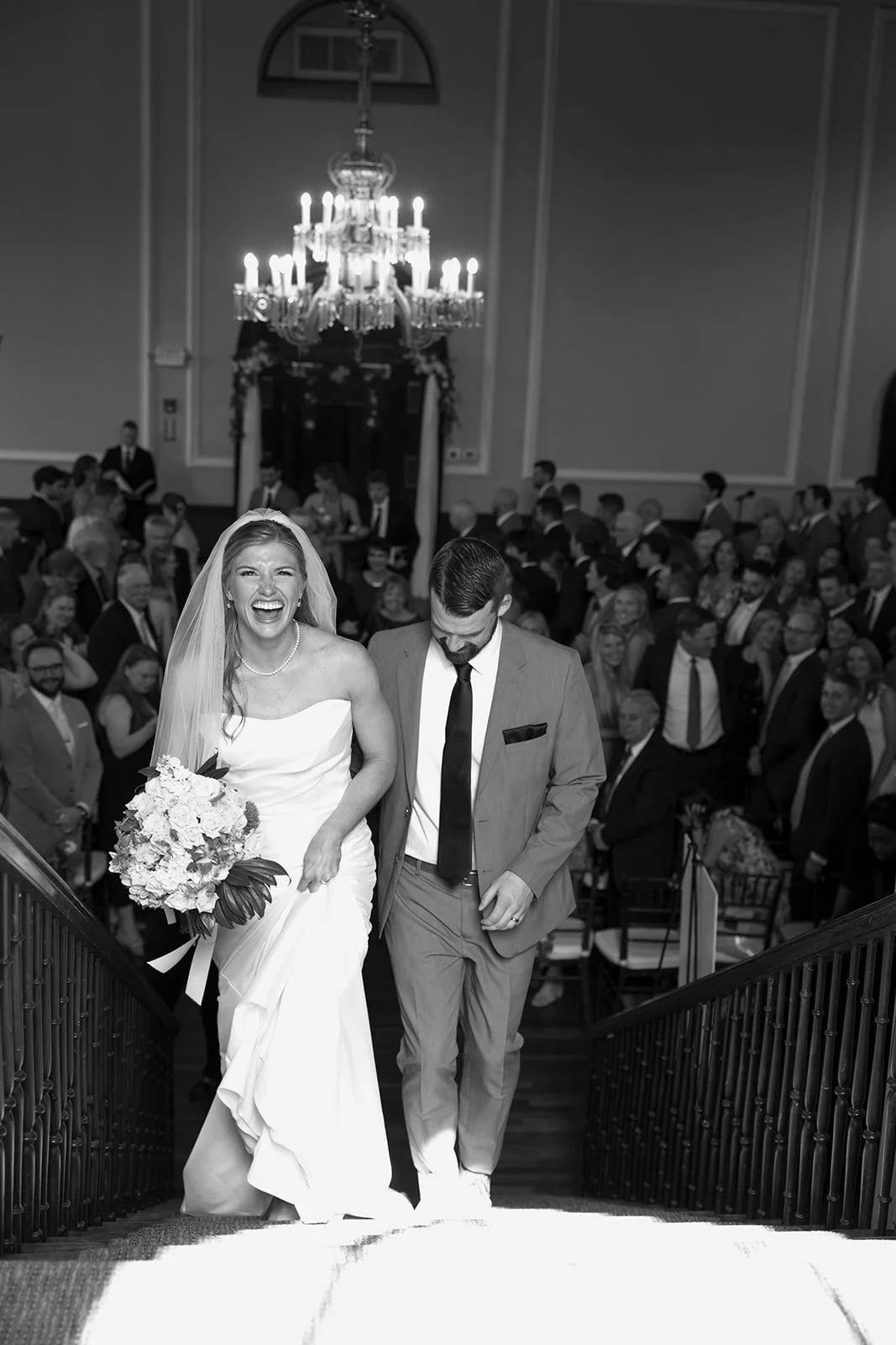 A joyful bride in a white wedding dress holding a bouquet of flowers, walking down the stairs with her groom in a suit, surrounded by wedding guests in an elegant, grand hall with a large chandelier overhead.